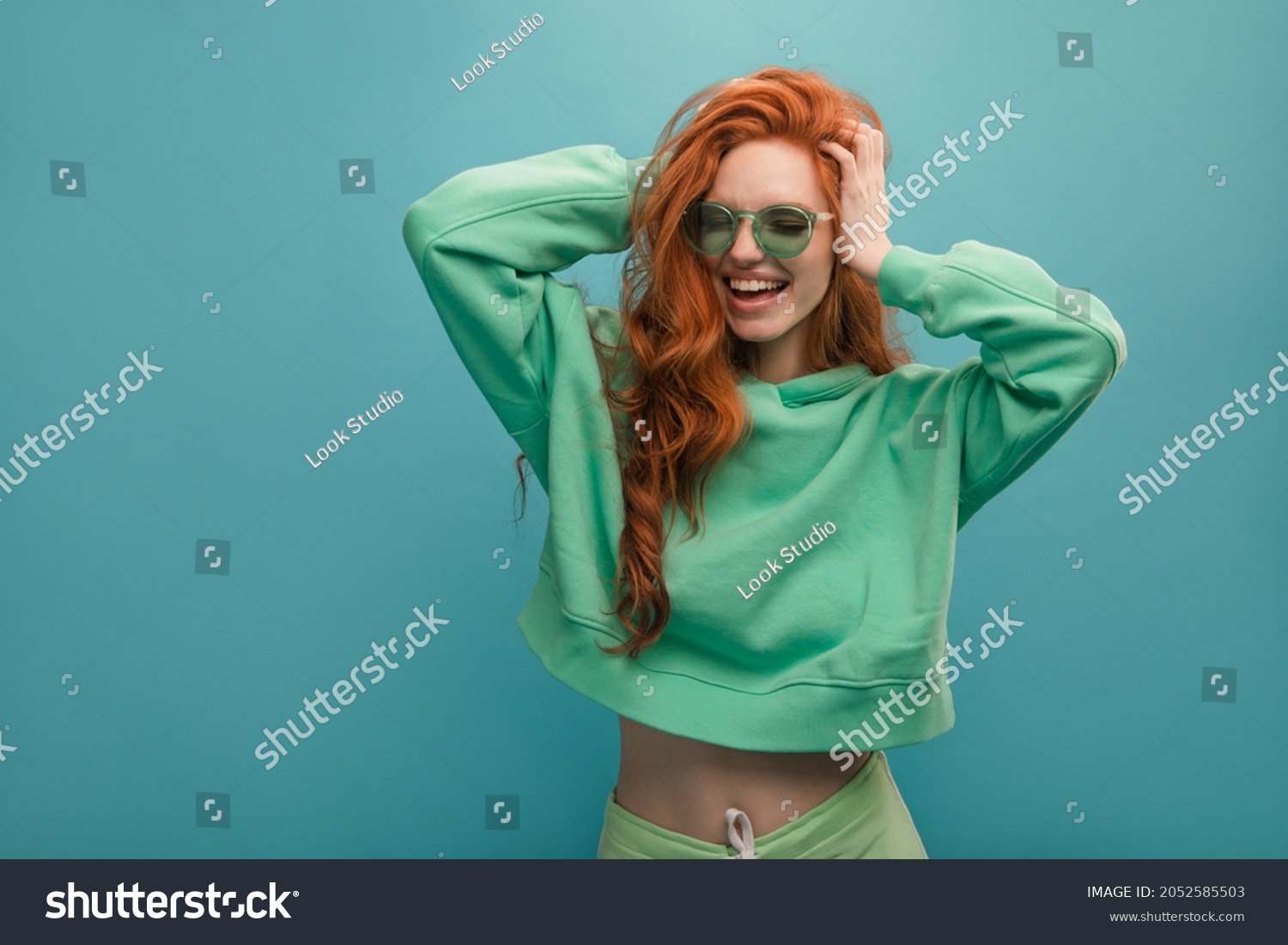 Disheveled young woman of European appearance is holding her head against blue background. Red-haired hipster in translucent glasses is wearing green hoodie. Concept of good mood, emotion #2052585503