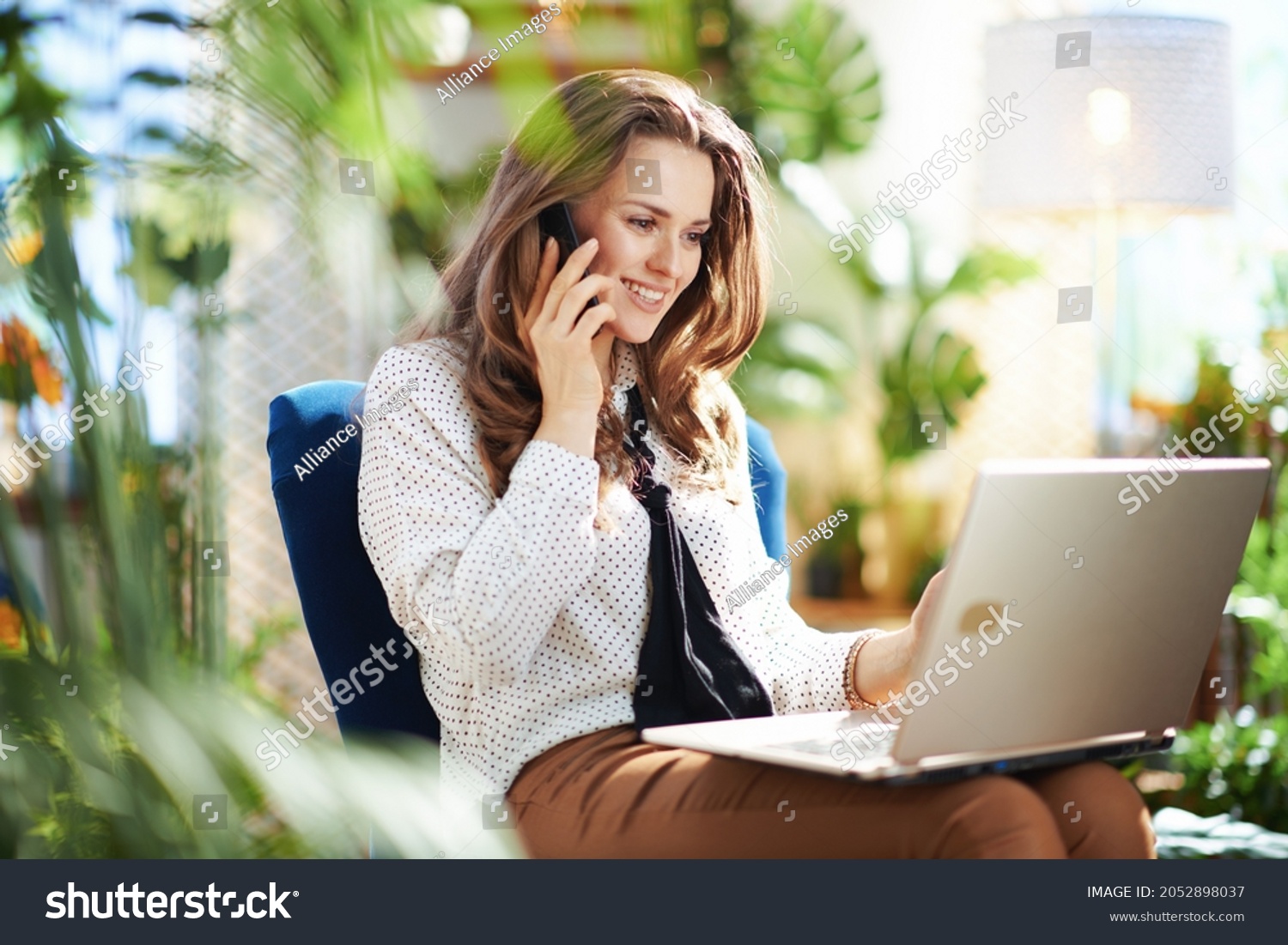 Green Home. happy modern 40 years old woman with long wavy hair in the modern house in sunny day using a smartphone and using laptop while sitting in a blue armchair. #2052898037