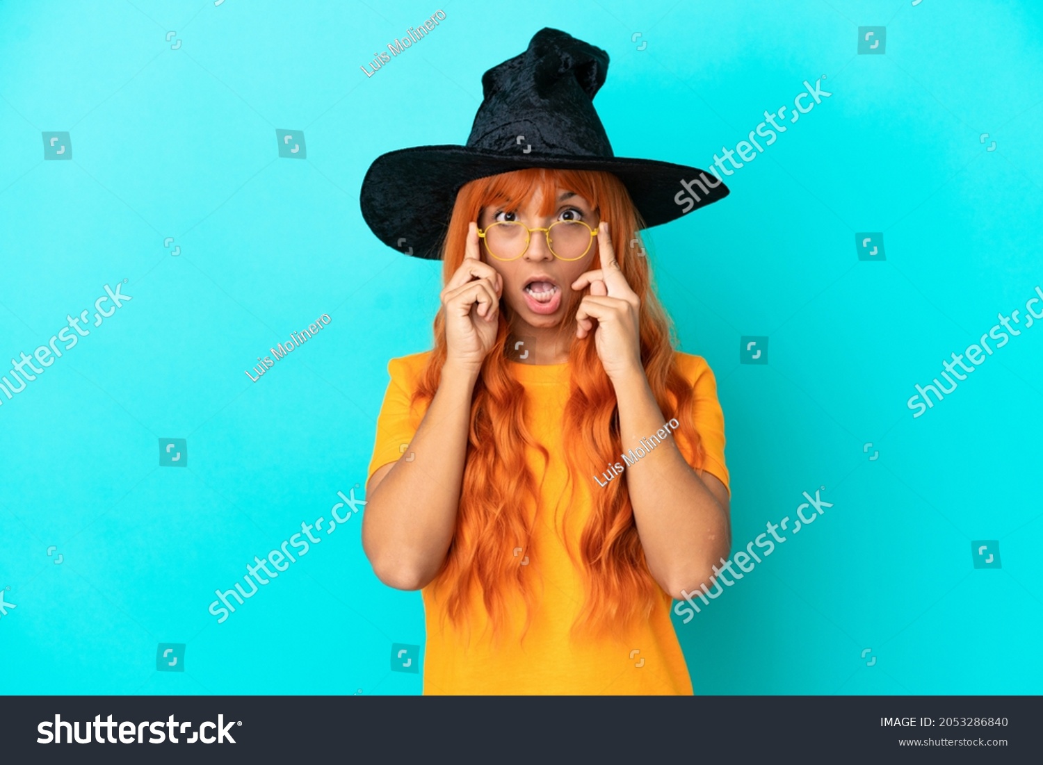 Young woman disguised as witch isolated on blue background with glasses and surprised #2053286840