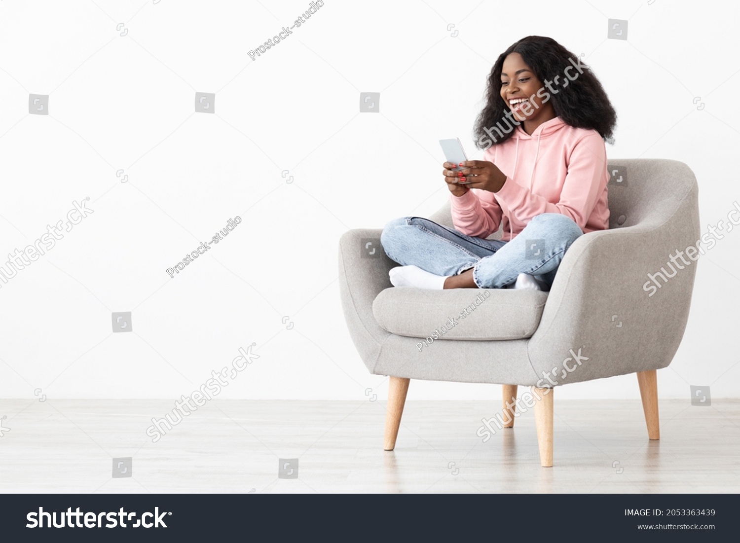 Cheerful african american young lady sitting in arm chair at home, using nice mobile application on brand new smartphone, shopping online, white background, panorama with copy space #2053363439