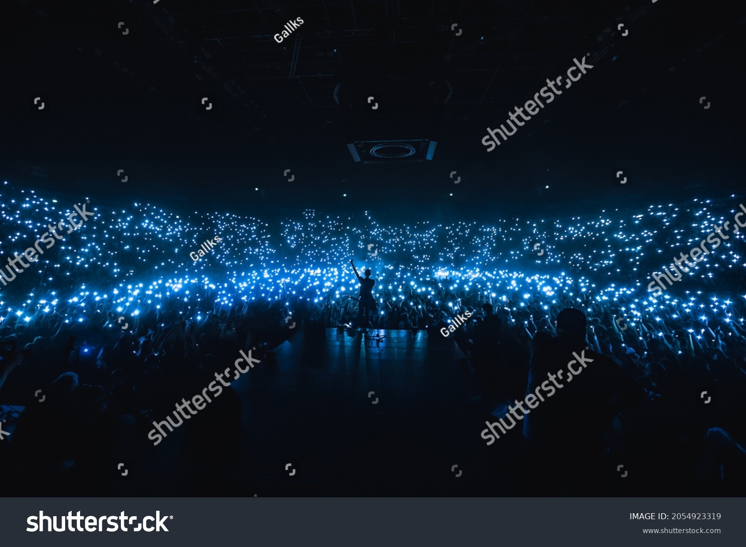 Vocalist in front of crowd on scene in stadium. Bright stage lighting, crowded dance floor. Phone lights at concert. Band blue silhouette crowd. People with cell phone lights. #2054923319