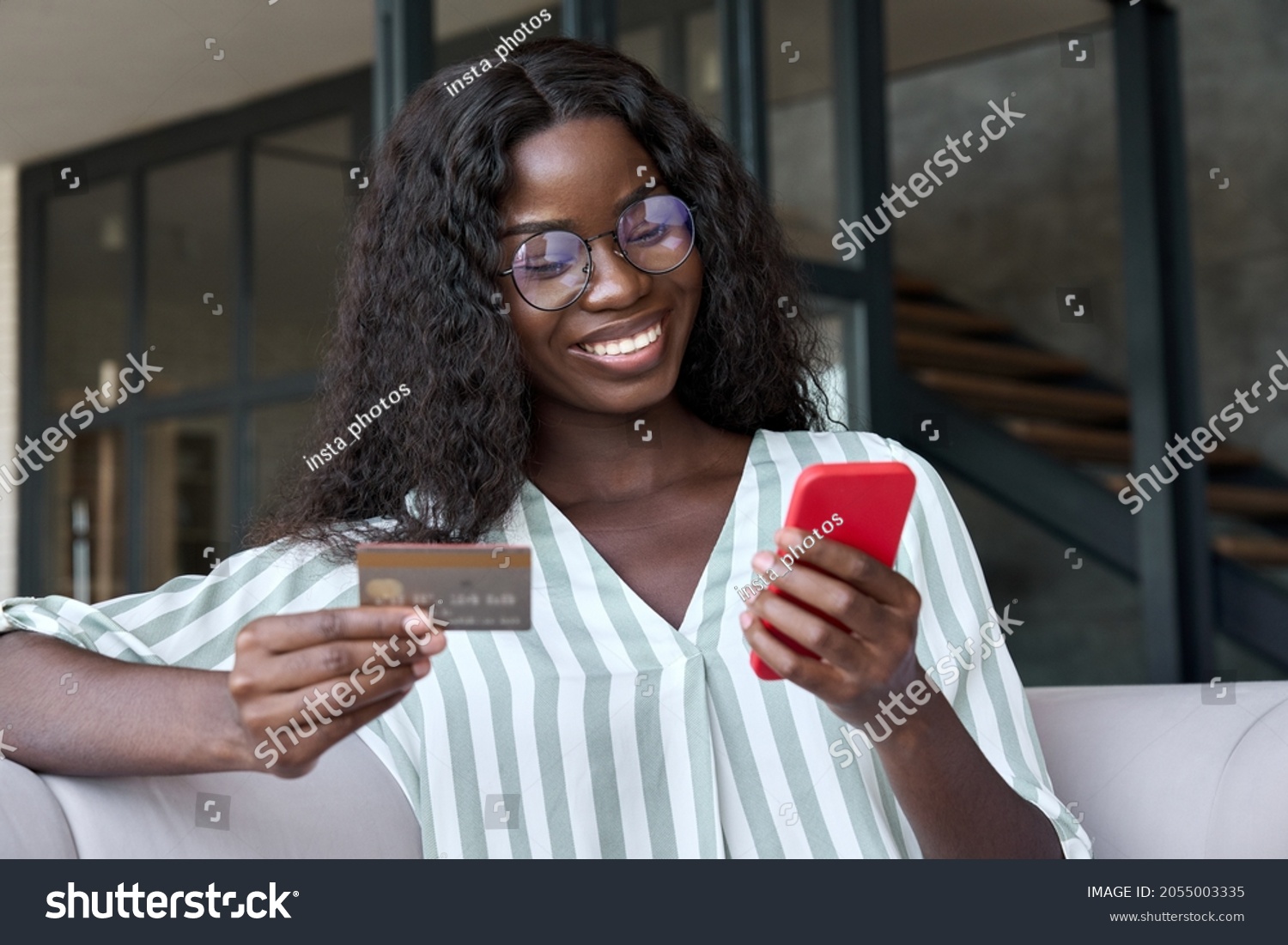 Happy young black African woman customer shopper holding credit card using cell phone mobile app buying fashion clothes paying online making purchase in ecommerce digital store on smartphone at home. #2055003335