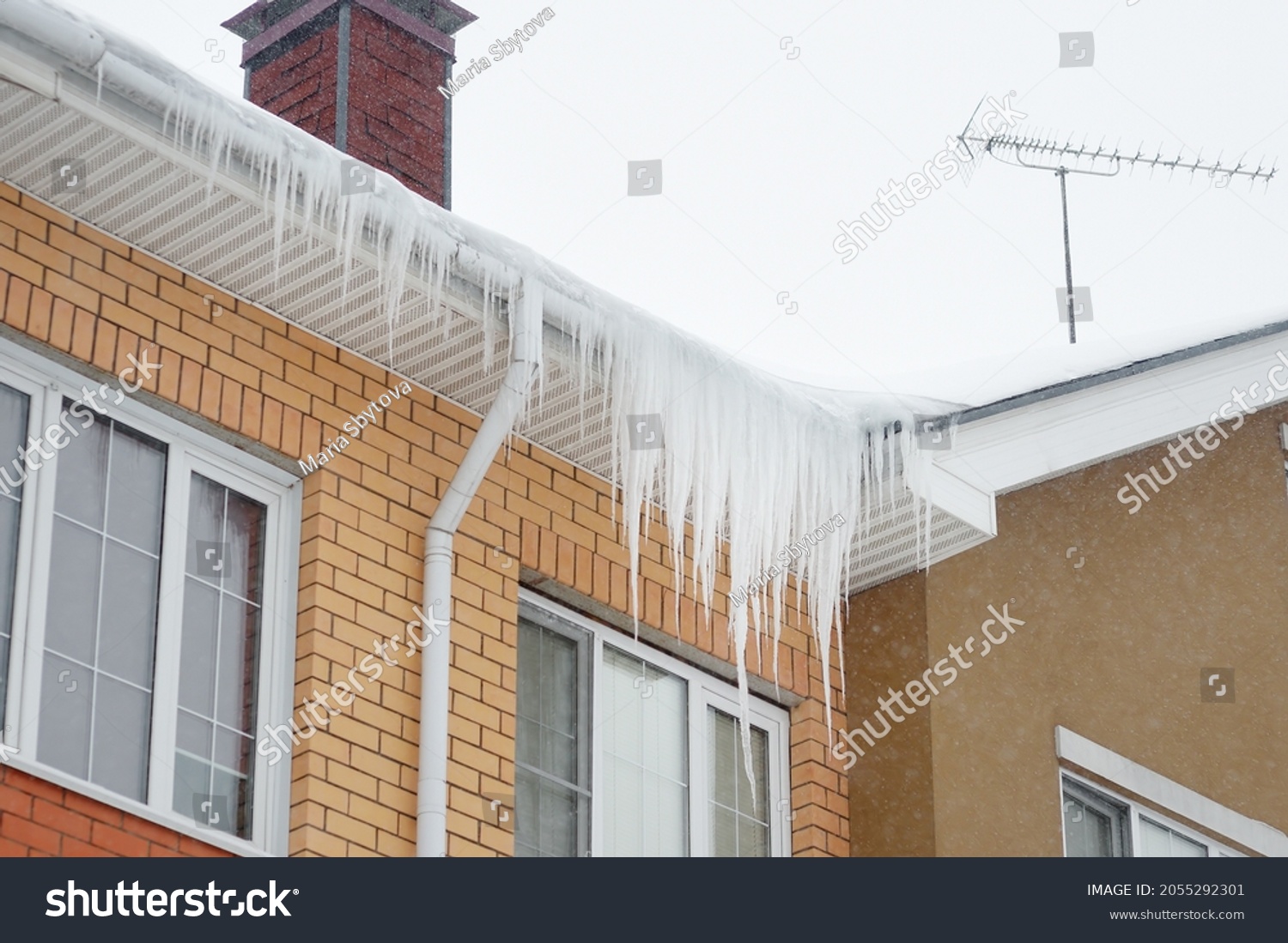 Big icicles on the roof of a townhouse on a snowy winter day among thaw. Cleaning the roofing from snow and icicles. #2055292301