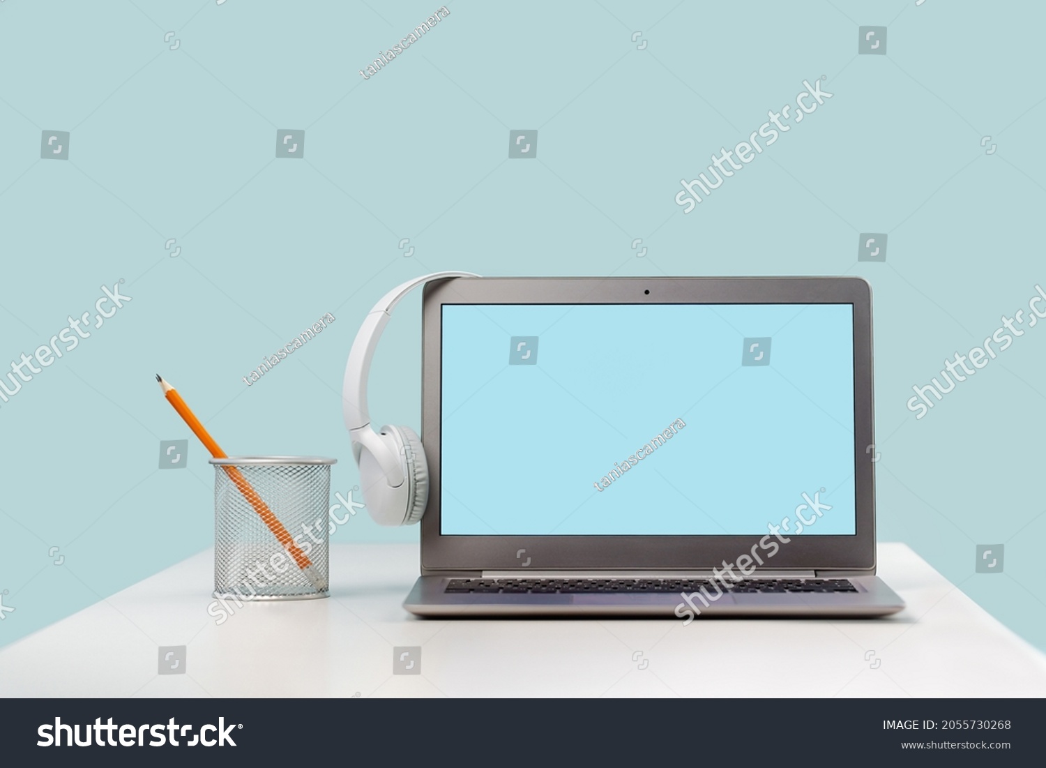 Front view of laptop mockup screen with wireless headphones on office desk. Pencils. Blue background. Distant learning. working from home, online courses or support. podcast. Helpdesk or call center #2055730268