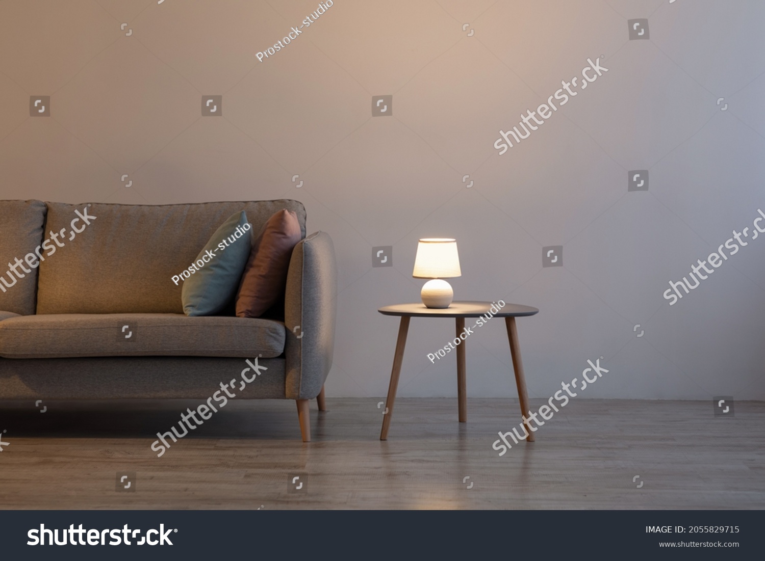 Couch with pillows, glowing lamp on table at home on gray wall background in evening in living room. Advertising of modern minimalist interior, comfortable furniture and real estate blog, free space #2055829715