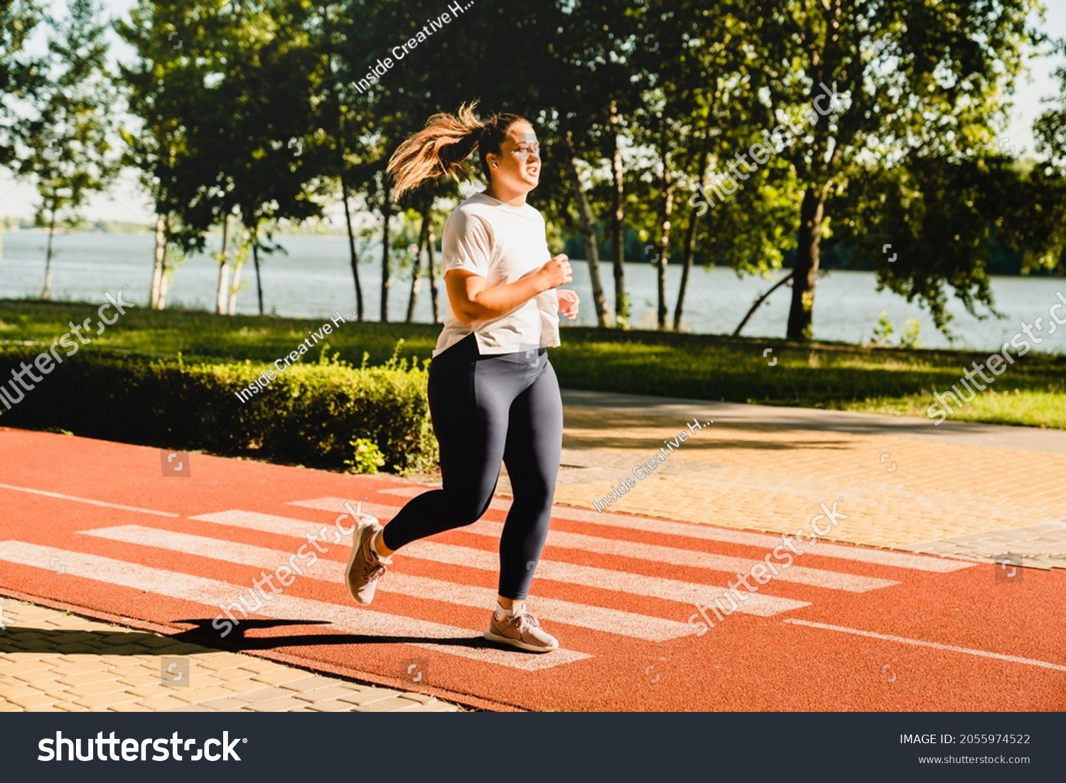 Full size portrait of a body positive plus-size plump woman athlete jogging running in fitness outfit, losing weight while listening to music in stadium outdoors #2055974522