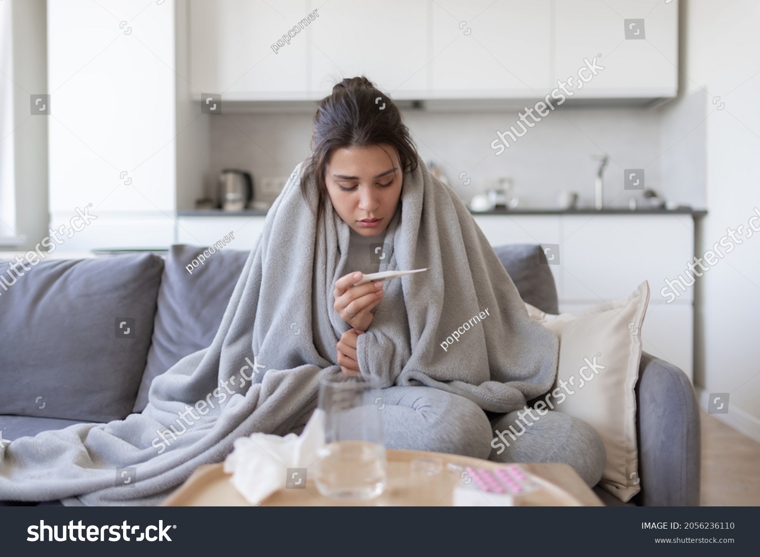Unhealthy upset young female with fever looking at thermometer with high degree temperature. Sick ill girl suffer from flu or cold, have covid-19 symptoms sitting covered blanket on couch at home. #2056236110