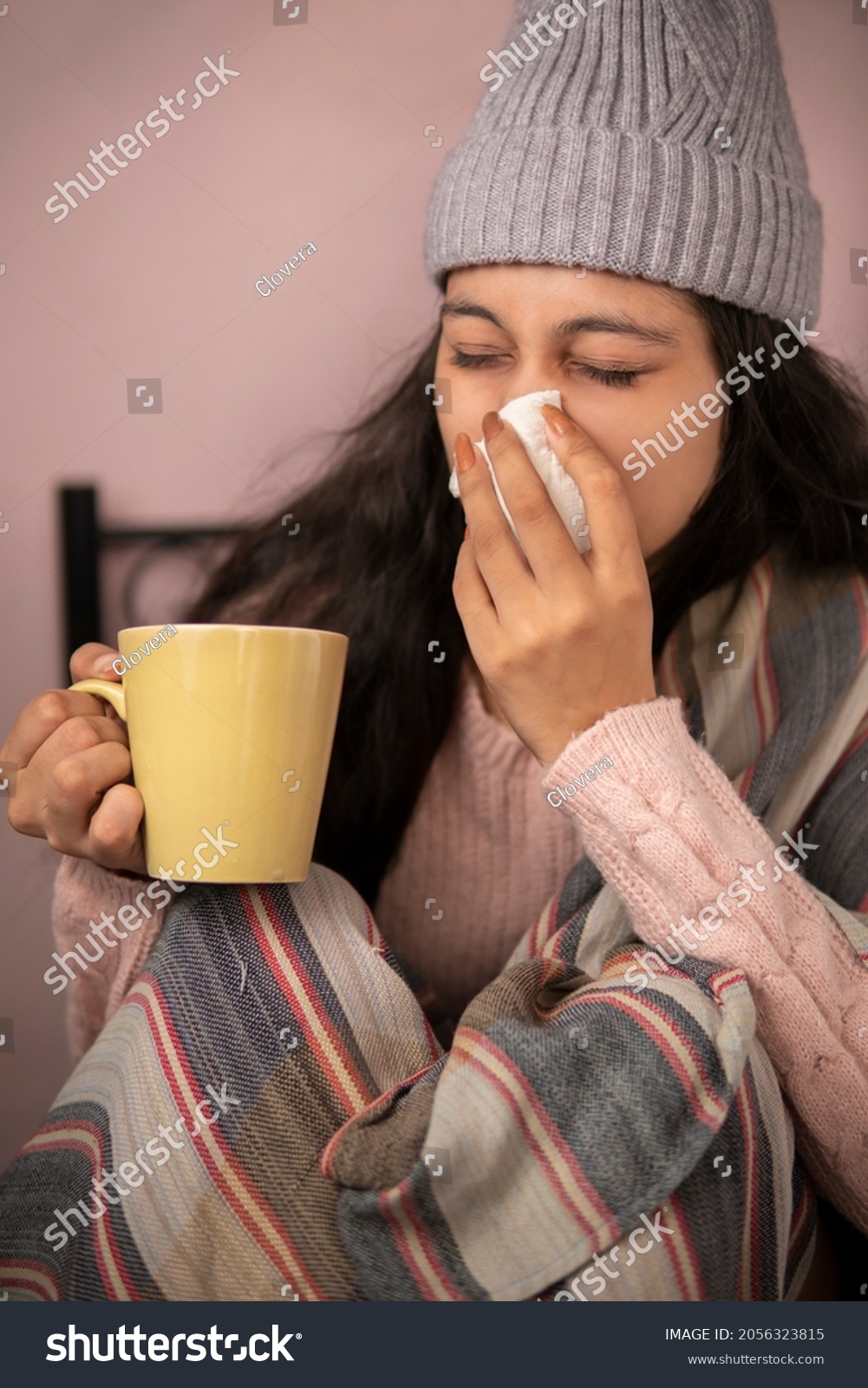 indoor image of a young woman getting sick with flu and cold and using a tissue paper to sneeze and blowing her nose while holding a coffee mug at home in winters. She is wearing warm clothes. #2056323815