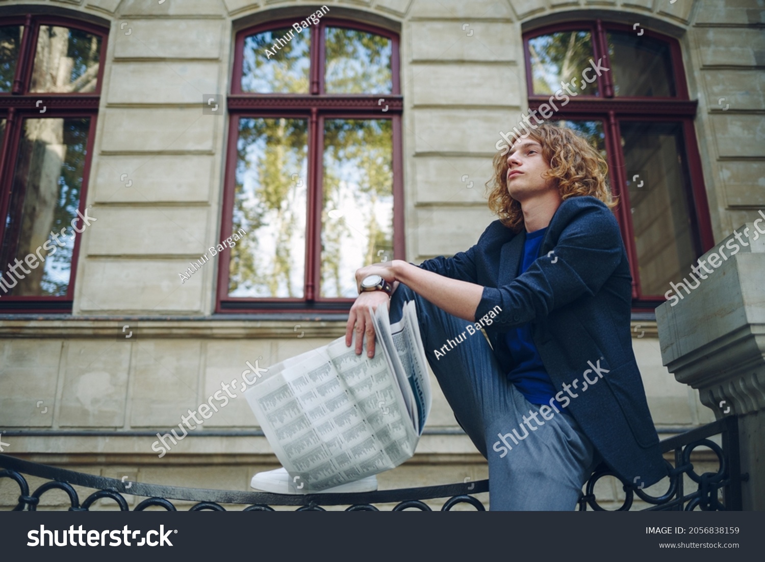 Young reddish, red haired attractive man with curly hair, holding a newspaper sitting near an urban old style building. Youth in action young leader thinking about future, city news. #2056838159