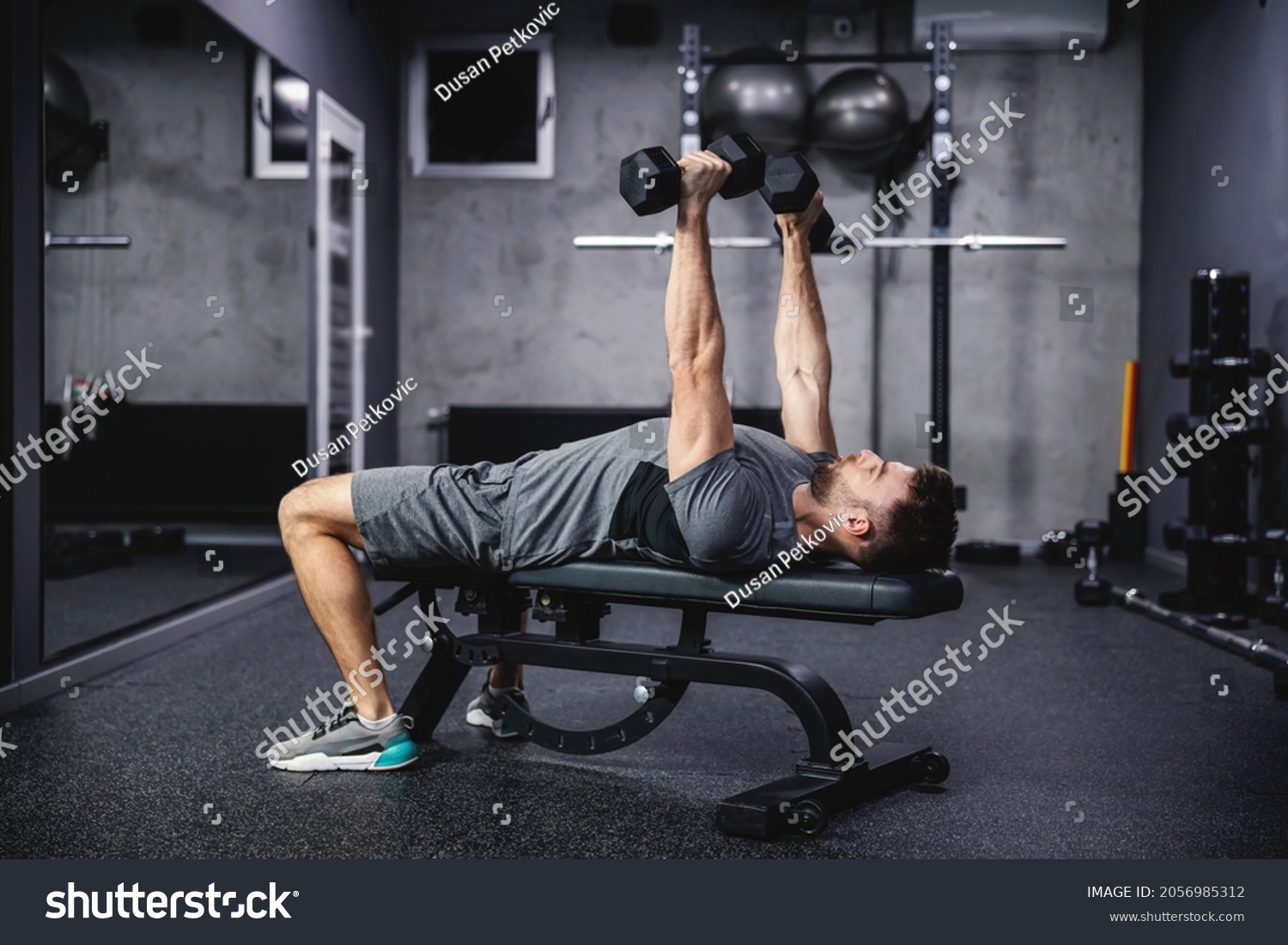 Chest and arm workout at sports bench with dumbbells. A man in sportswear lies on a sports bench and lifts dumbbells, training with a load. Strength, powerful body and physical endurance #2056985312