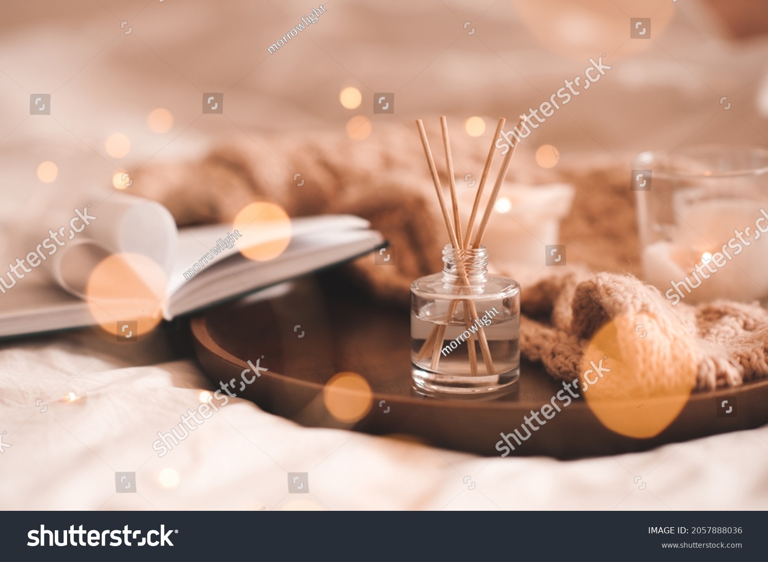 Home perfume in glass bottle with wood sticks, scented burn candles, open paper book and knit wool textile on ray in bedroom close up. Aromatherapy cozy atmosphere lifestyle. Winter warm xmas season.  #2057888036