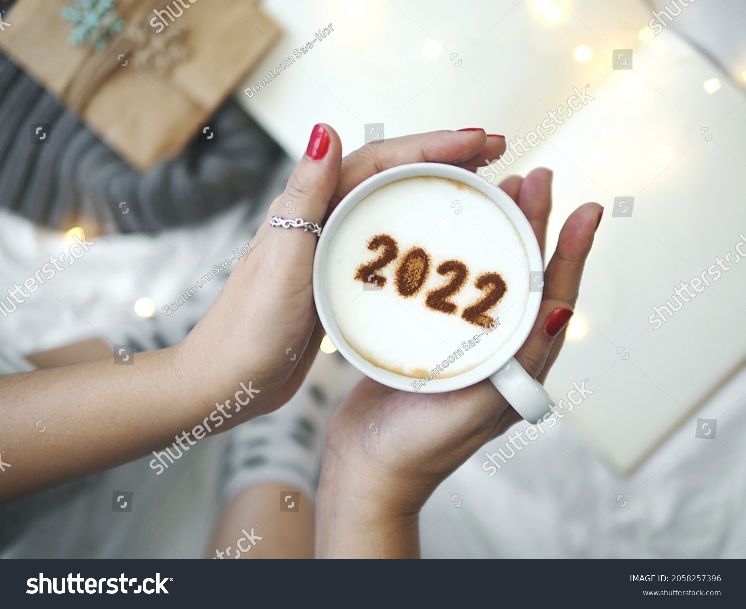 Number 2022 on frothy surface of cappuccino served in white cup holding by female hands on blurred white bed with string lights. New year new you, Holidays food art theme Happy New Year. (top view) #2058257396