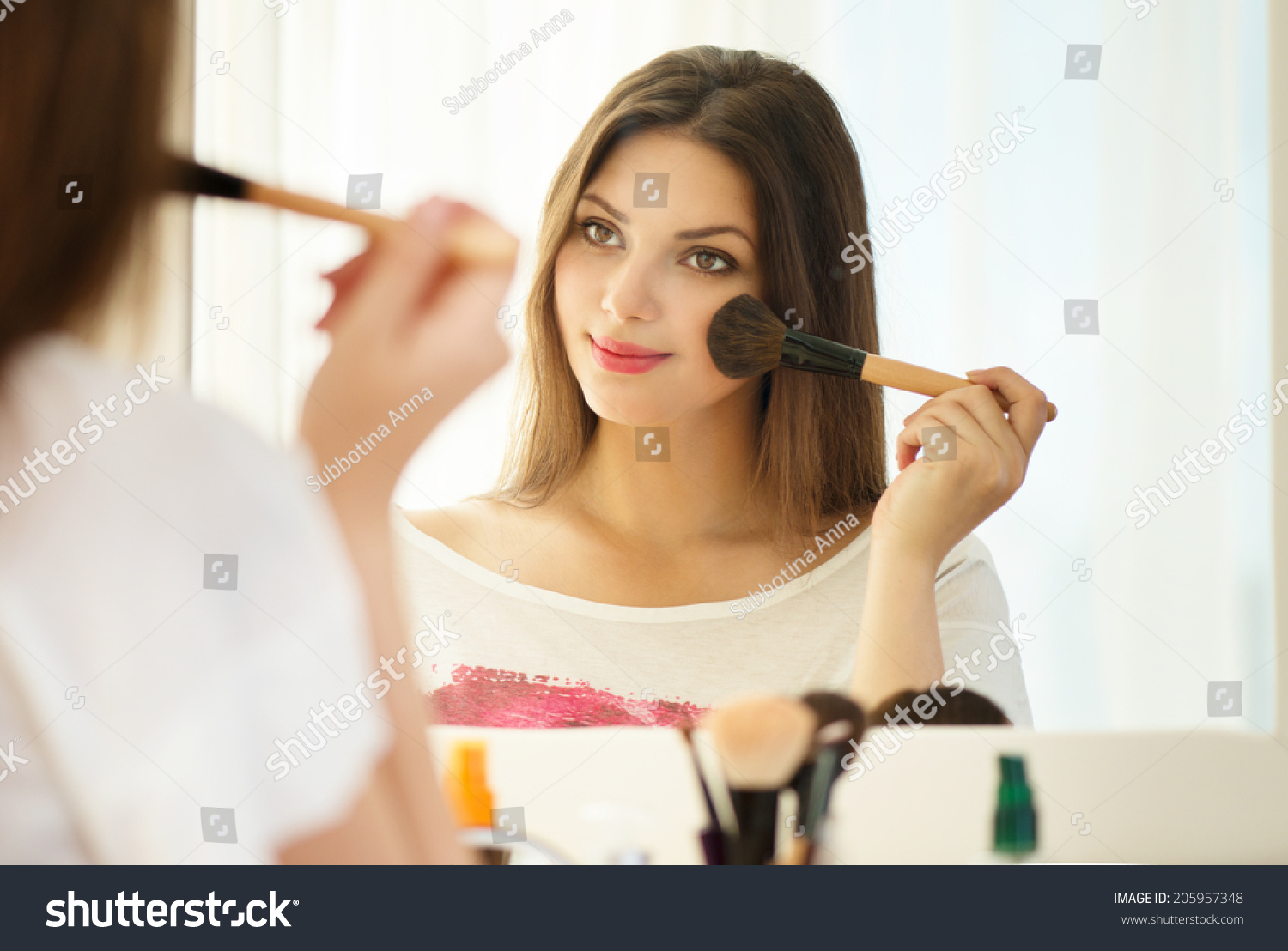 Beauty woman applying makeup. Beautiful girl looking in the mirror and applying cosmetic with a big brush. Girl gets blush on the cheekbones. Powder, rouge  #205957348