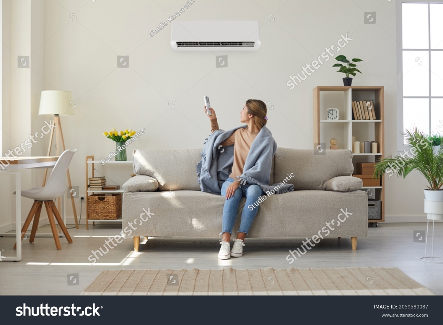 Woman who's sitting on sofa under warm plaid in living room switches off her air conditioner on wall. Young girl adjusting modern AC system, regulating temperature and enjoying cool fresh air at home #2059580087