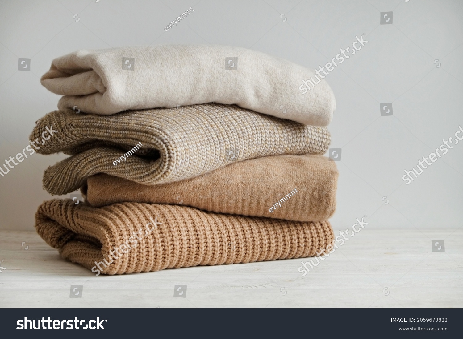 Bunch of knitted warm pastel color sweaters with different knitting patterns stacked in messy pile on white wooden table, white wall background. Fall winter season knitwear. Close up, copy space. #2059673822