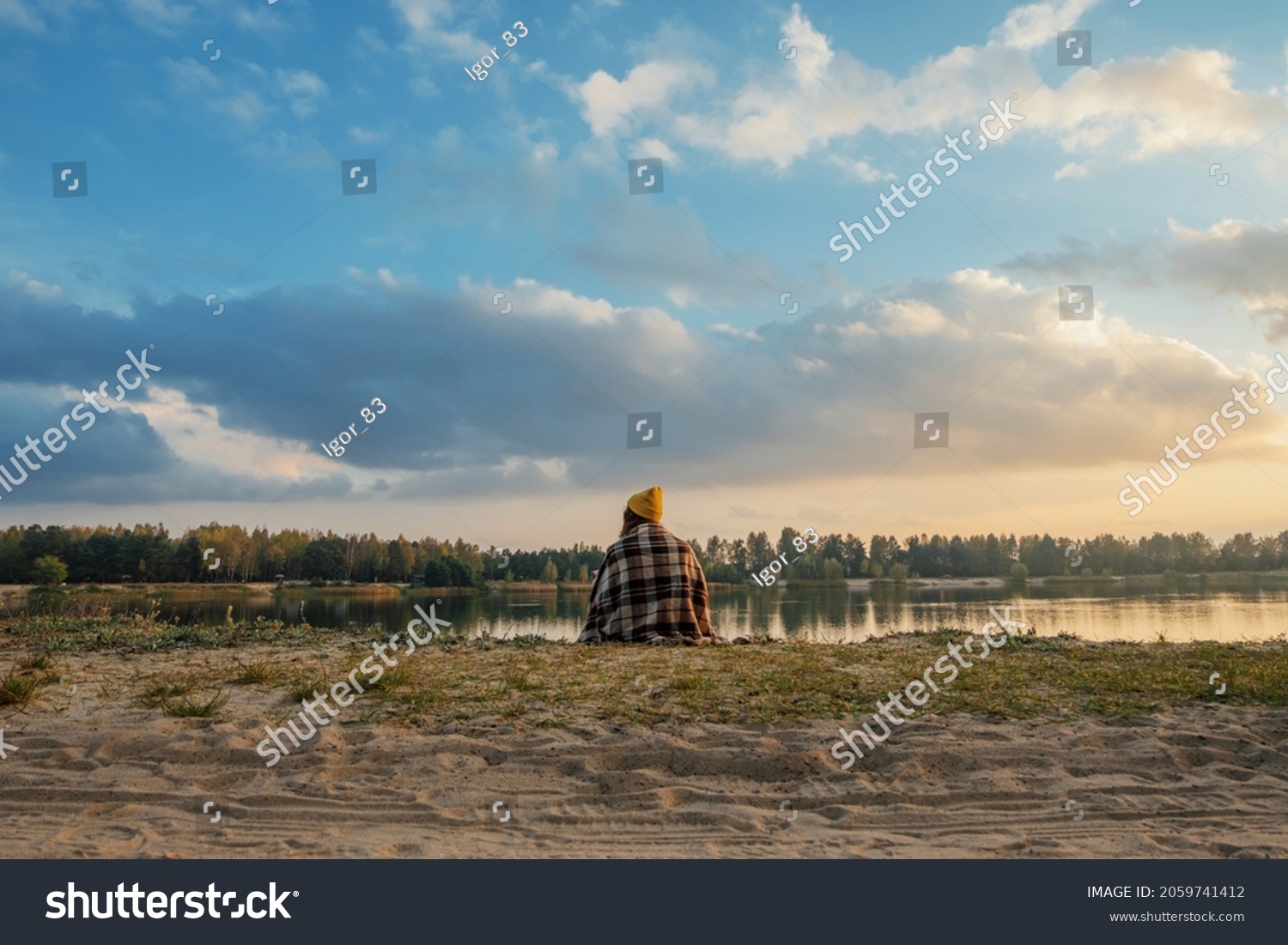  Girl traveler in autumn sits in a plaid blanket on the shore and looks at the sunset #2059741412