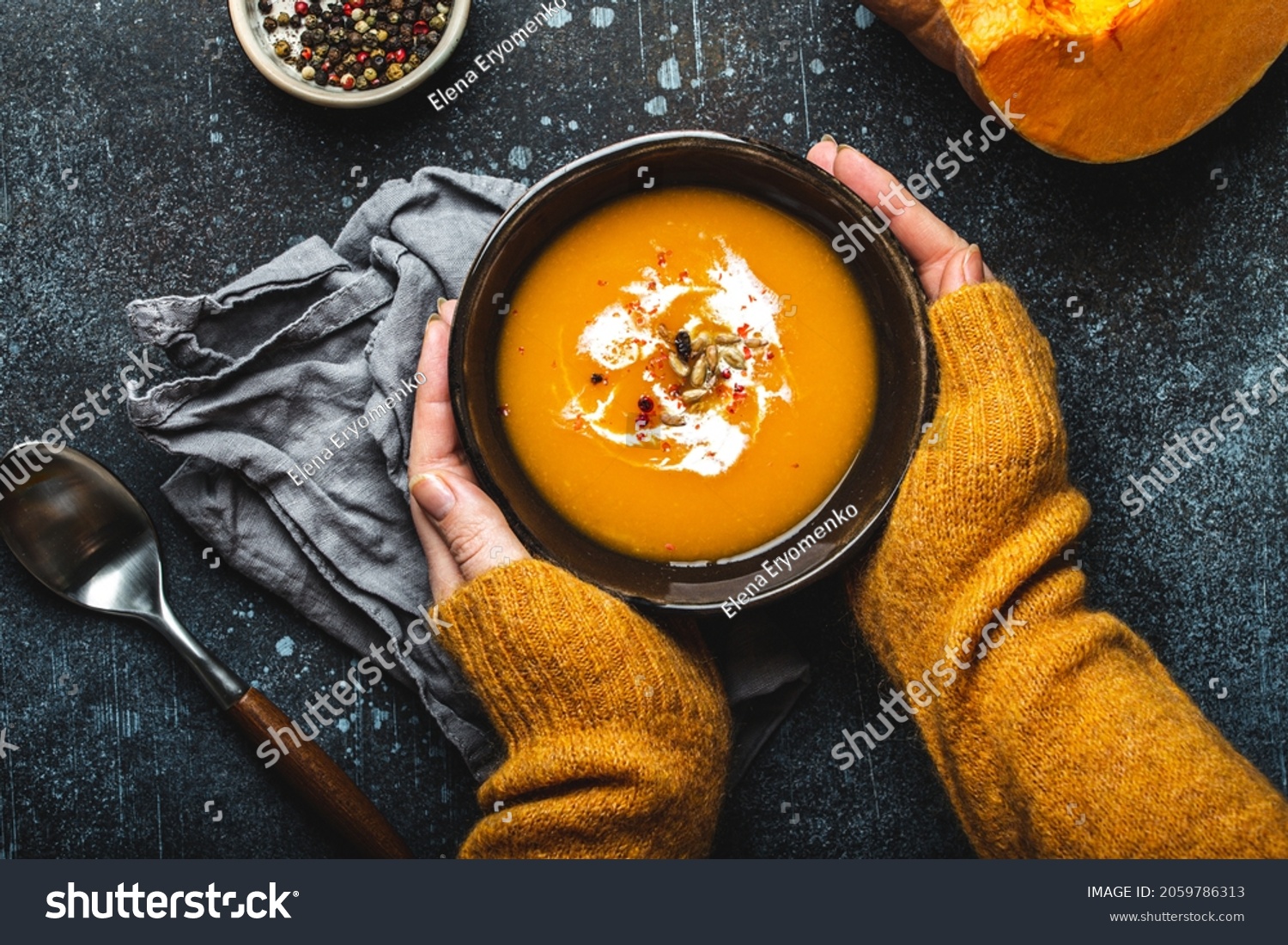 Female hands in yellow knitted sweater holding a bowl with pumpkin cream soup on dark stone background with spoon decorated with cut fresh pumpkin, top view. Autumn cozy dinner concept 
 #2059786313