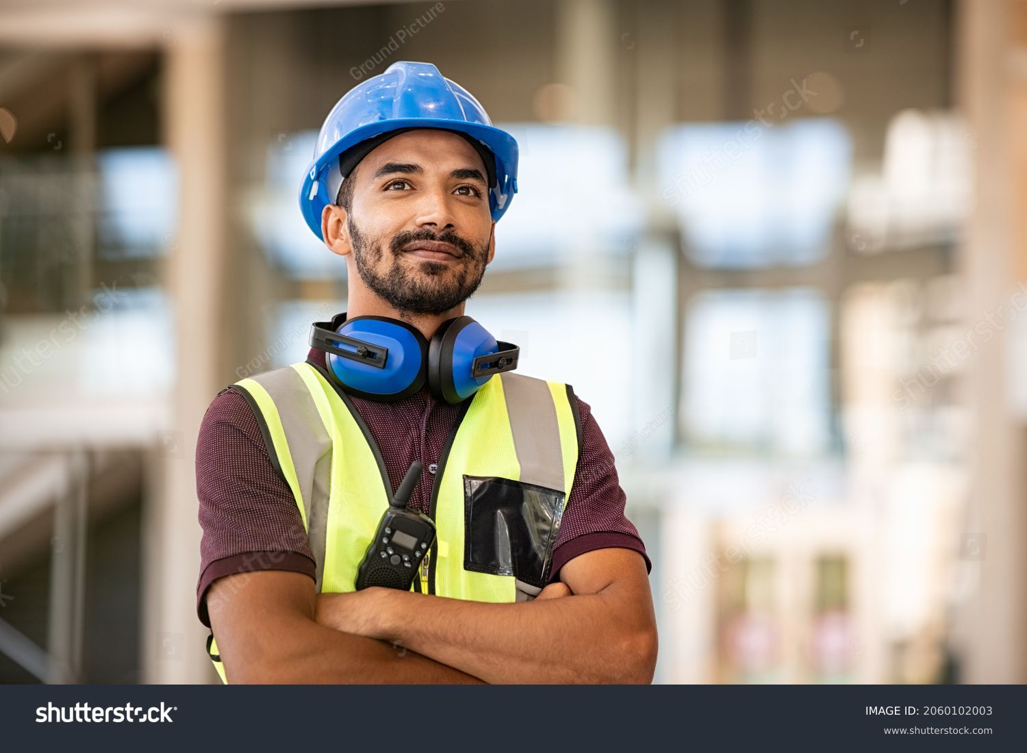 Indian construction site manager standing with folded arms wearing safety vest and helmet, thinking at construction site. Portrait of mixed race manual worker or architect with satisfaction. #2060102003