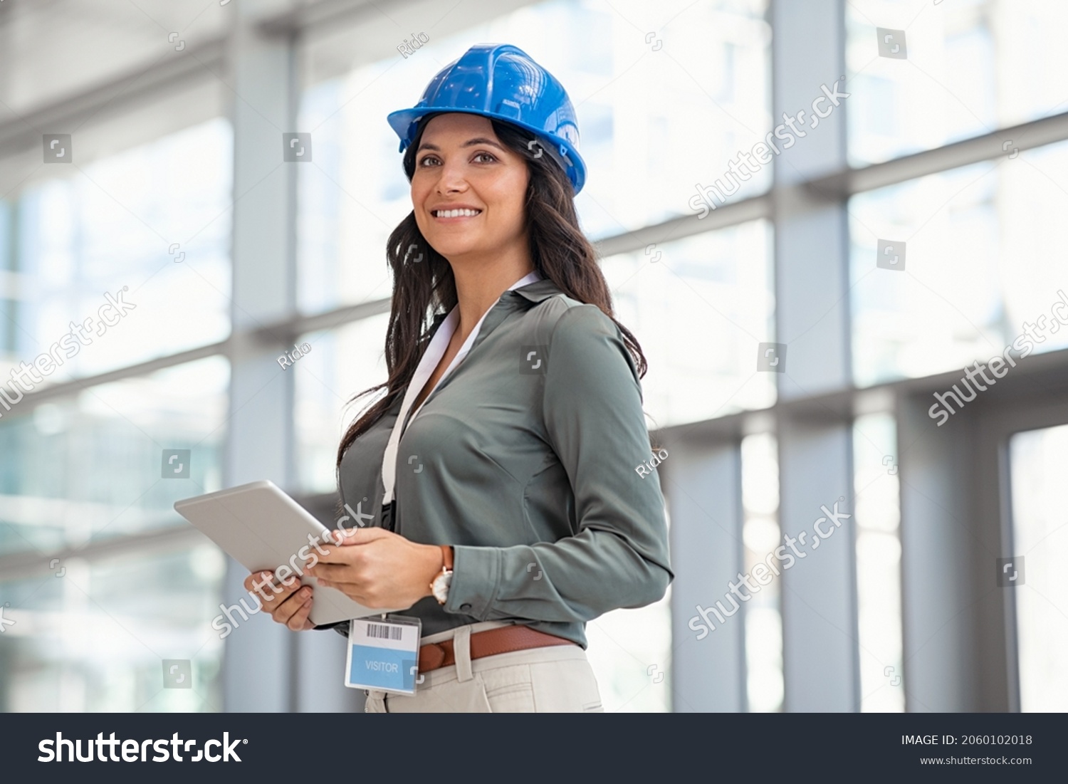 Mid adult woman architect wearing hardhat at construction site while working on digital tablet. Supervisor wearing safety helmet while working in a building site. Successful and proud inspector. #2060102018