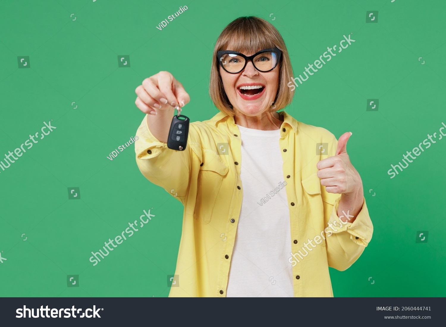 Elderly smiling happy caucasian woman 50s in glasses yellow shirt hold car keys fob keyless system show thumb up gesture isolated on plain green background studio portrait People lifestyle concept. #2060444741