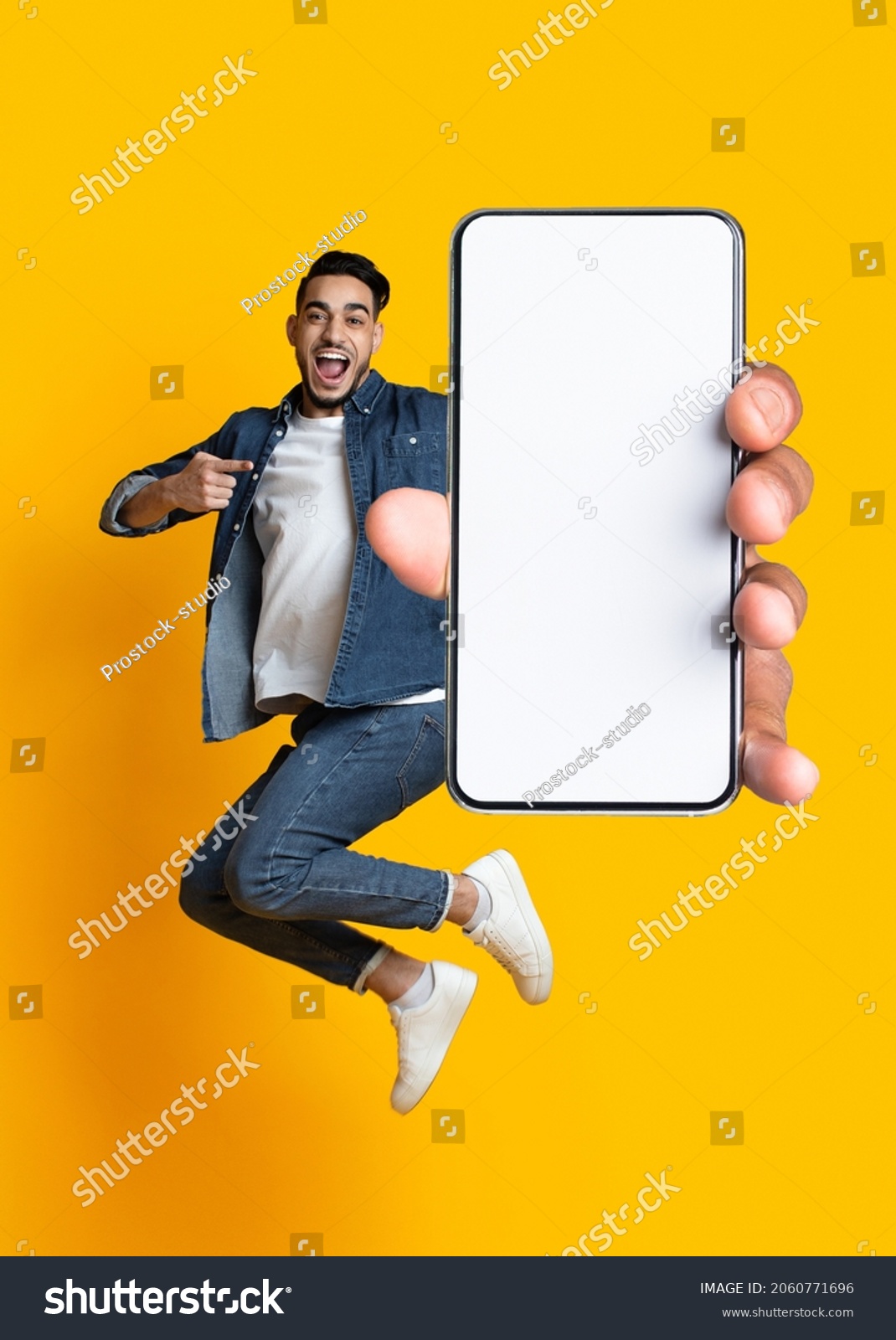 Handsome young Arab guy jumping on air, pointing at cellphone with empty screen on orange studio background. Millennial Muslim man demonstrating mockup for mobile app design #2060771696