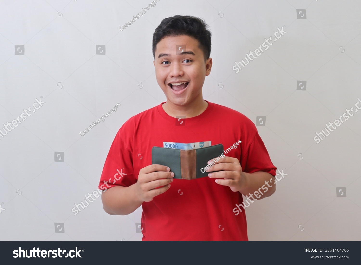 Portrait of attractive Asian man in red t-shirt looking at camera, holding a wallet with rupiah banknote. Isolated image on gray background #2061404765