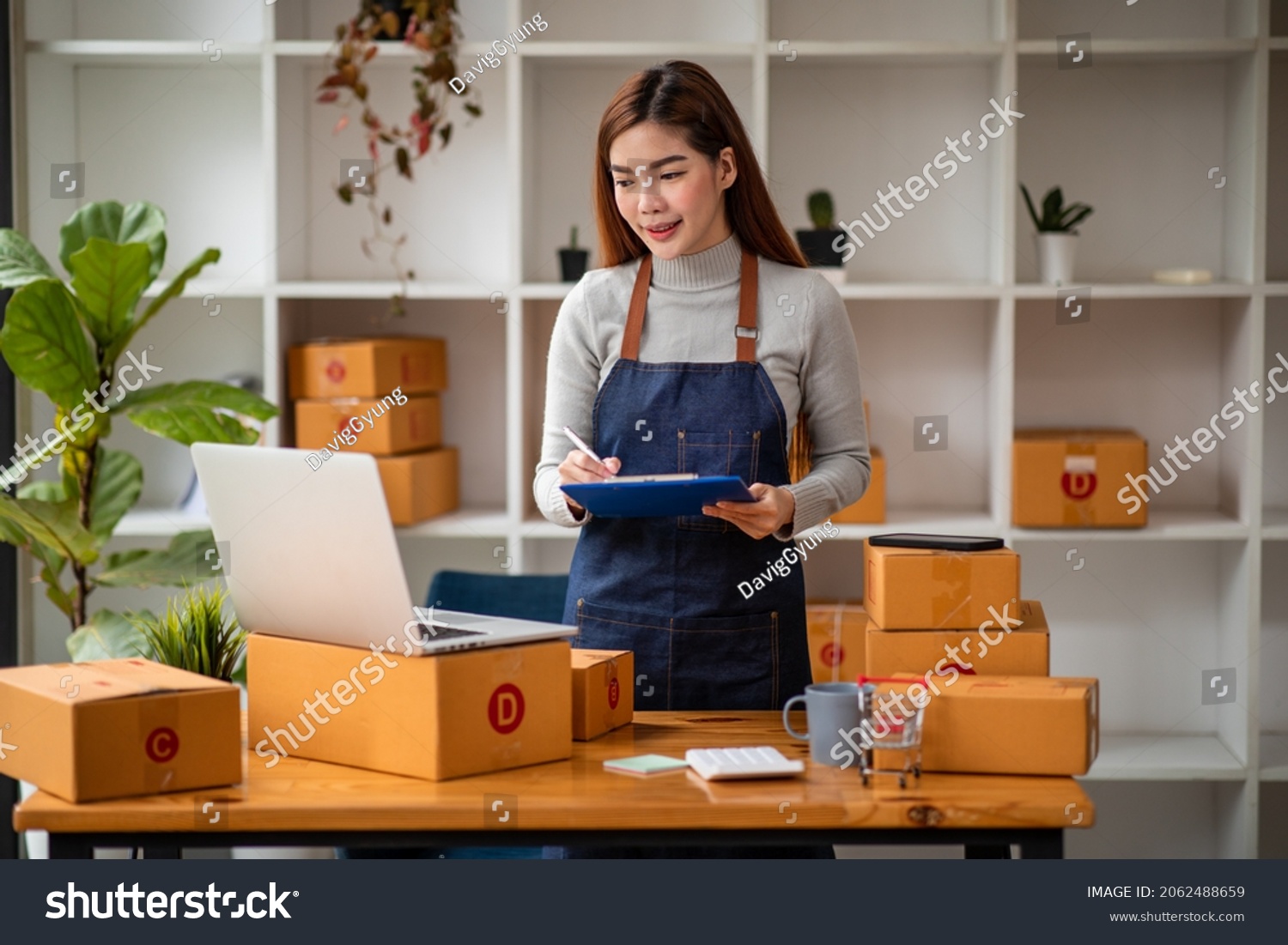 Stylish small business owner Young Asian woman using laptop computer in her home office, small business entrepreneur SME, working with a box at home, Online selling, e-commerce, packing concept. #2062488659