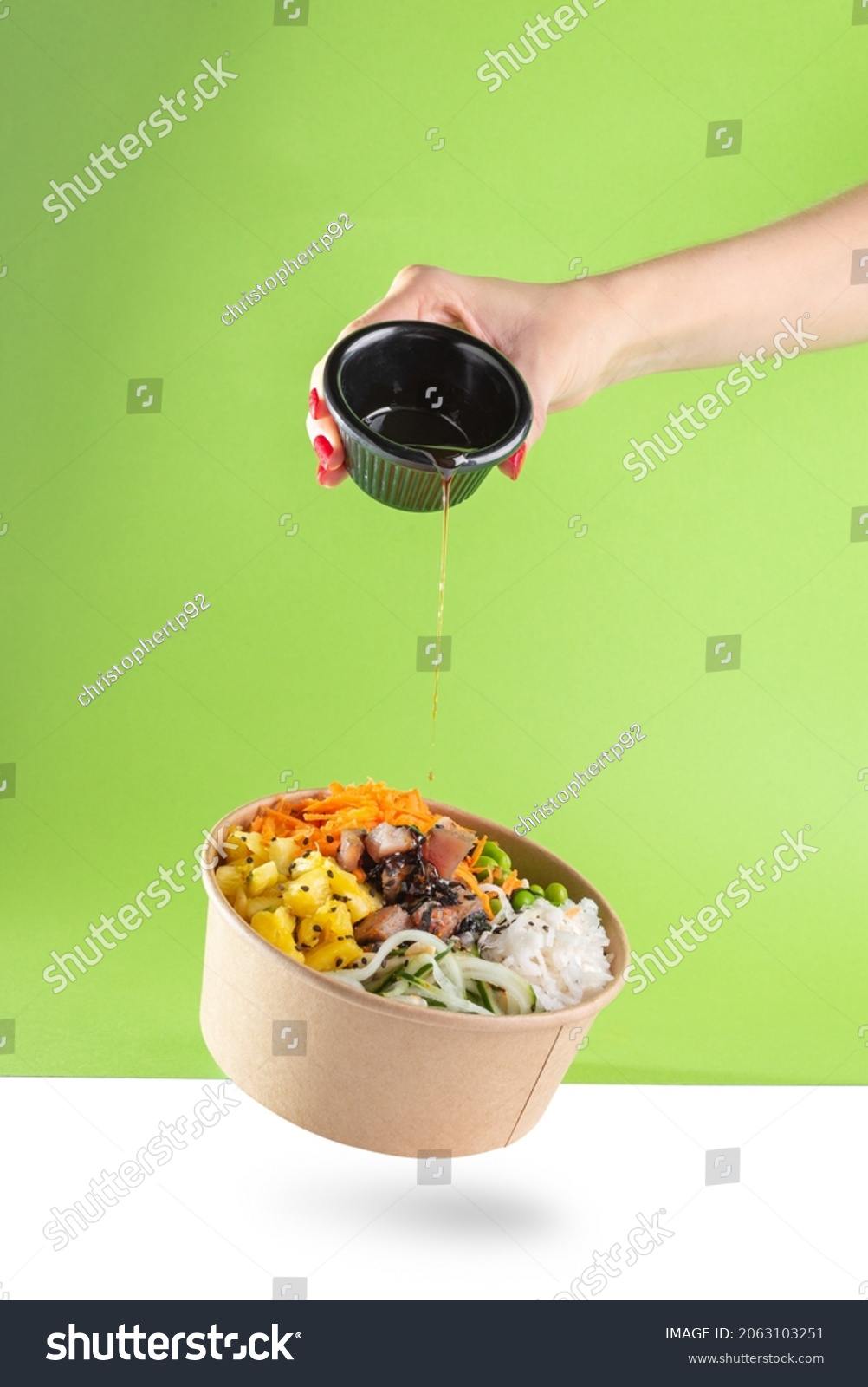 A woman's hand pours soy sauce into a floating bowl of salad poke and tuna, carrot, cucumber, and pineapple with a green background. white floor #2063103251