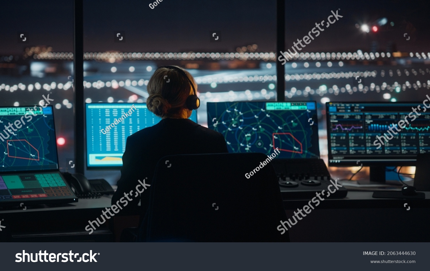 Female Air Traffic Controller with Headset Talk on a Call in Airport Tower at Night. Office Room is Full of Desktop Computer Displays with Navigation Screens, Airplane Flight Radar Data for the Team. #2063444630