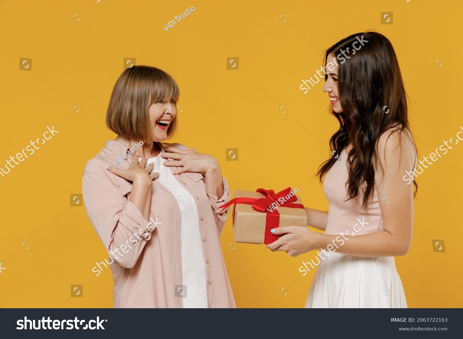 Side view two fun young smiling happy daughter mother together couple women in casual beige clothes gifting birthday present with red ribbon isolated on plain yellow color background studio portrait. #2063722163