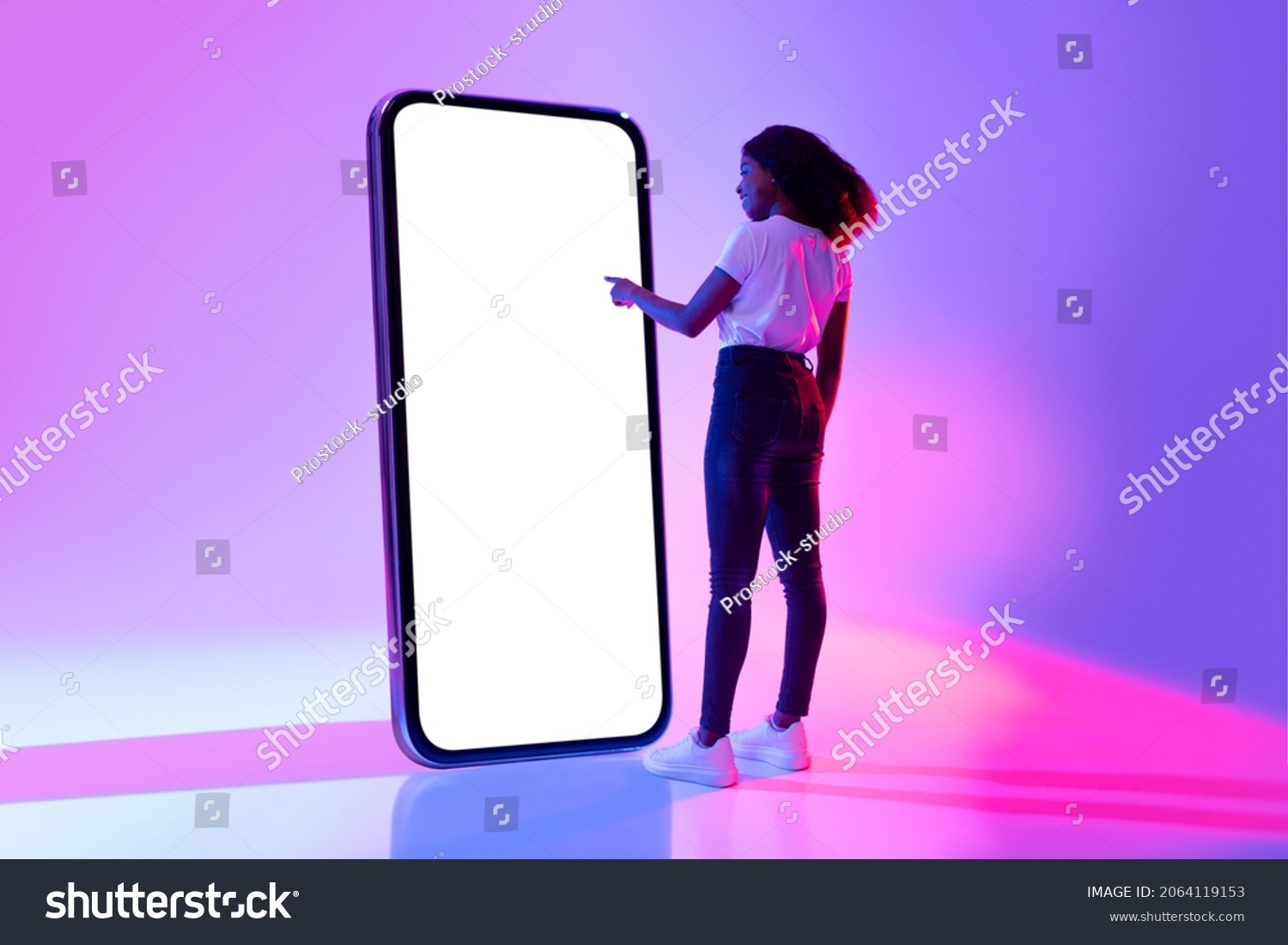 Full length of young black woman touching screen of giant smartphone in neon light, mockup for new mobile app or website design. African American lady interacting with big cellphone, space for ad #2064119153