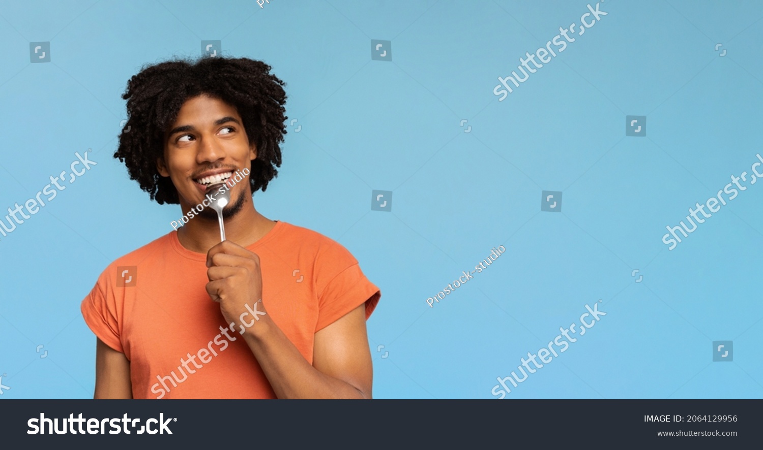Curious and hungry young african american man with spoon in his mouth looking at copy space, dreaming about delicious healthy food, choosing something to eat, blue studio background, panorama #2064129956