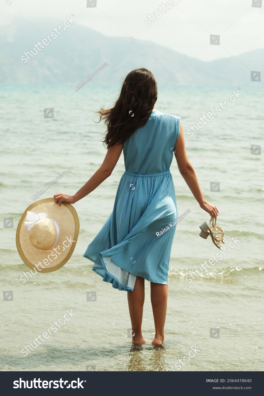 Image of a young brunette woman in a blue dress walking barefoot on a beach and dangles his feet in the water. Young woman in summer white dress with straw hat looking to a sky and sea. Back view. #2064478640
