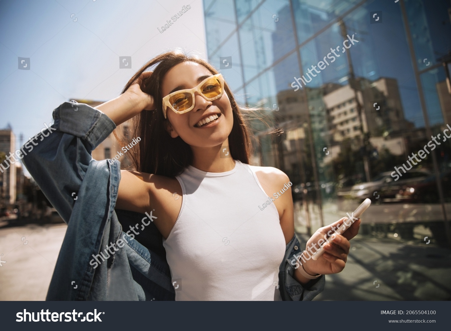 Close-up of fashionably dressed asian tourist in stylish glasses, eyes closed, walking relaxed in center of capital. Brunette in denim jacket with phone in hand. Lifestyle, female beauty concept #2065504100