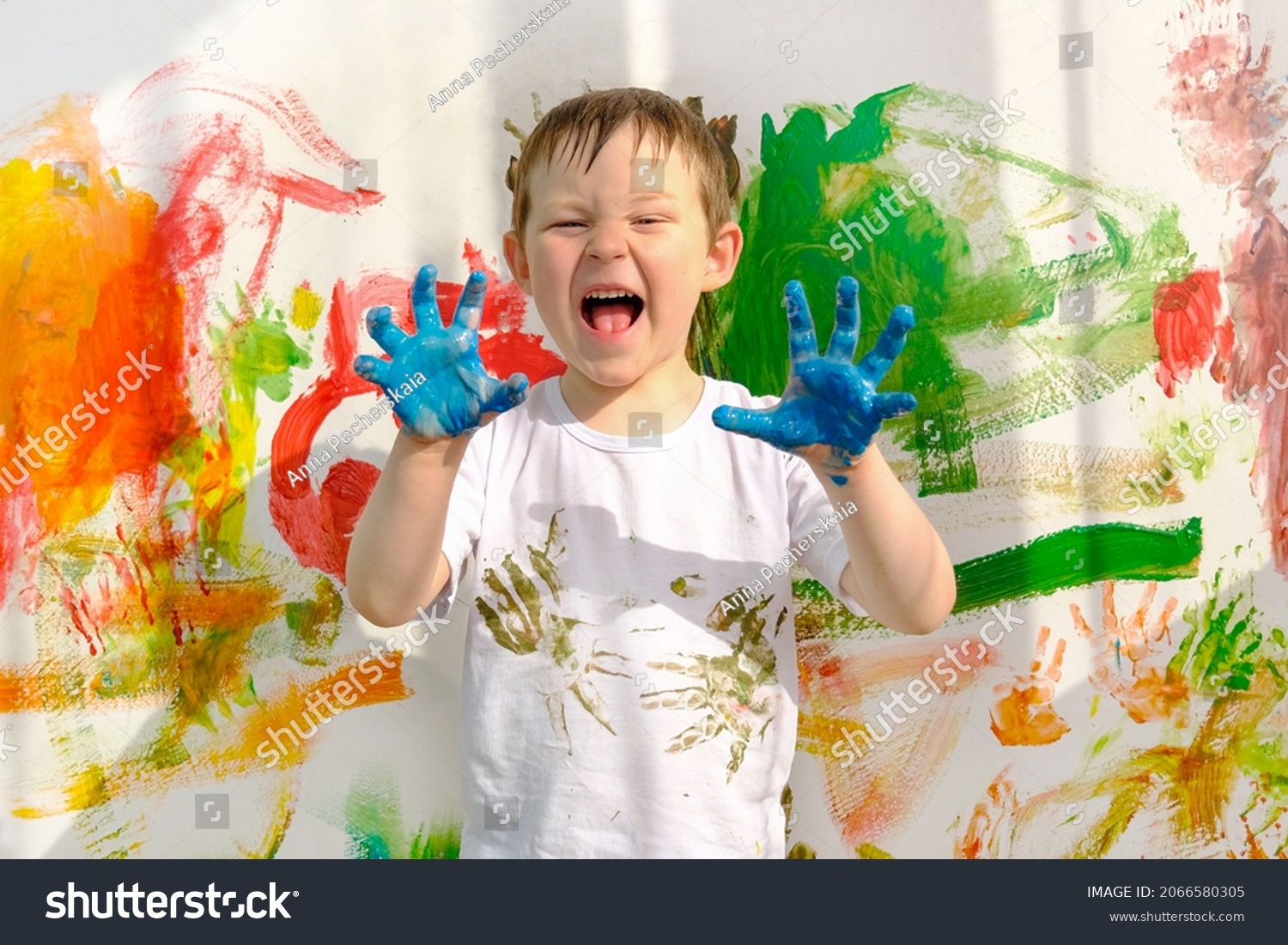 a boy in a white T-shirt, stained with paint, and dirty blue hands, stands against the background of a multi-colored wall. The child painted with his hands. A funny, lovable boy, looks into the frame. #2066580305