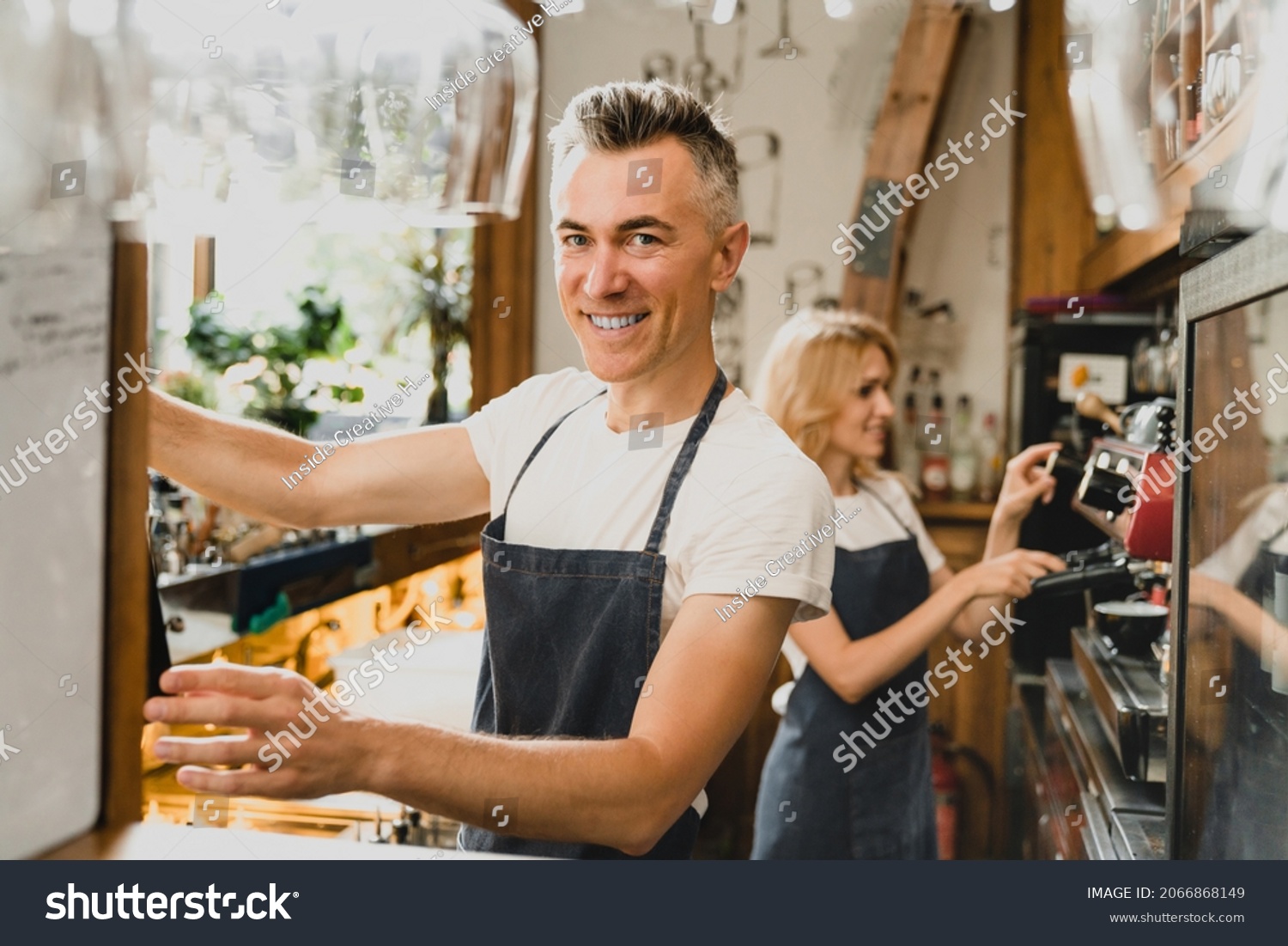 Confident smiling middle-aged small business owners barista bartenders waiters in blue aprons brewing coffee working preparing drinks orders for customers at the bar counter in restaurant cafe. #2066868149