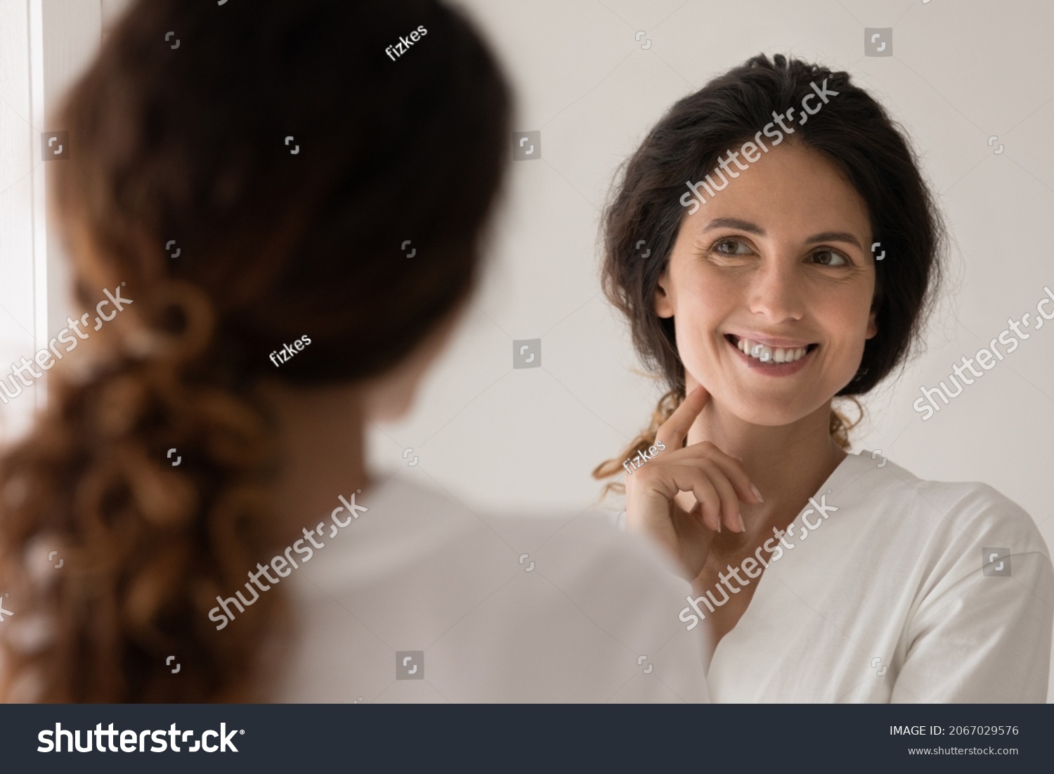 Beautiful Hispanic woman wear white bathrobe admire face view looking in mirror while standing in bathroom enjoy facial perfectness after skin procedure, applied cream, treatment for skincare beauty #2067029576
