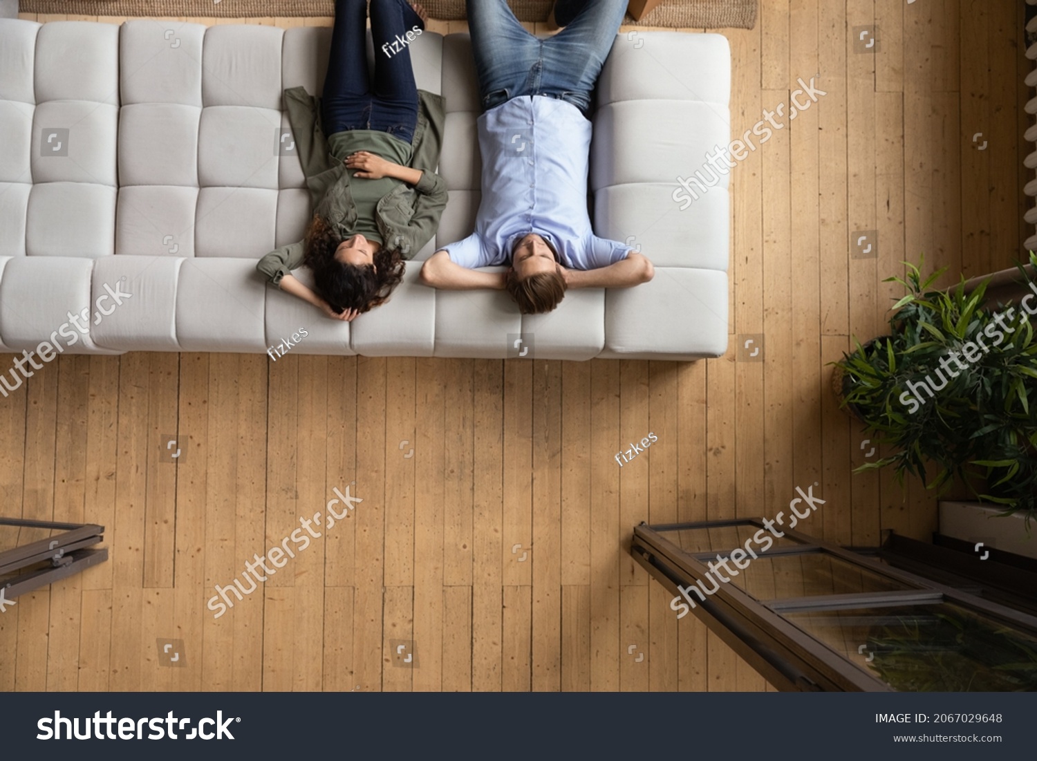 Serene couple put hands behind head relaxing on cozy sofa, above view. Homeowners family enjoy rest on fashionable furniture, breathing fresh conditioned air inside modern living room. Tenant concept #2067029648