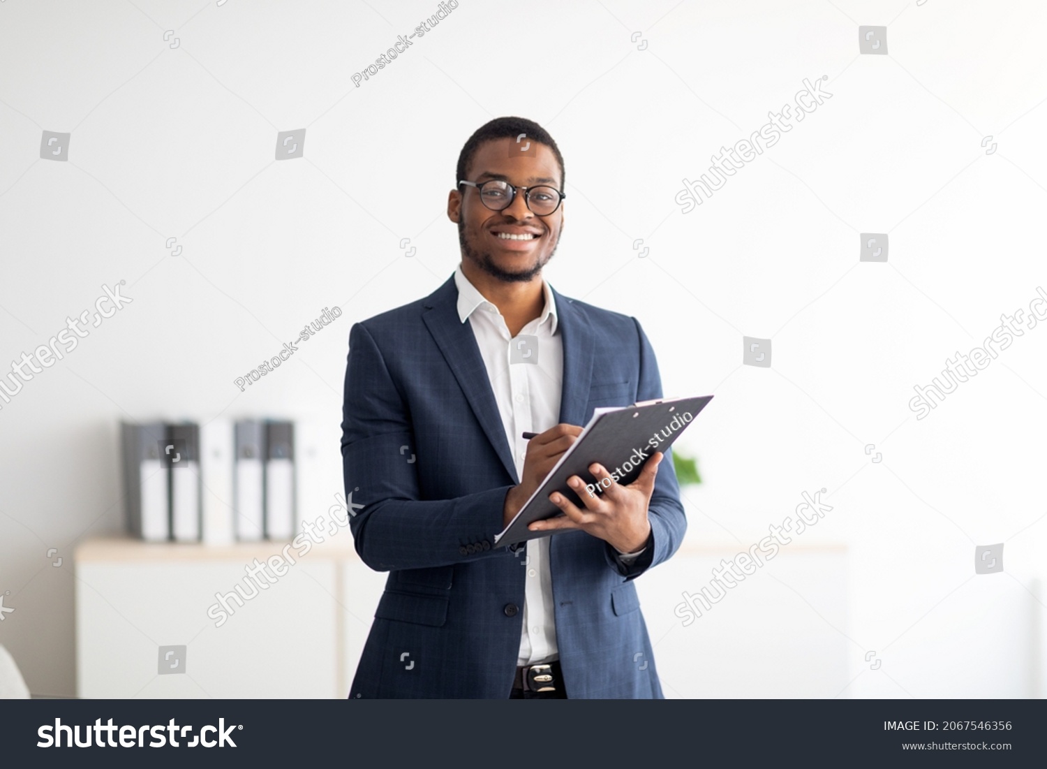Happy young black male psychologist writing in clipboard, looking at camera and smiling in modern office. Psychotherapy services, mental health professional concept #2067546356