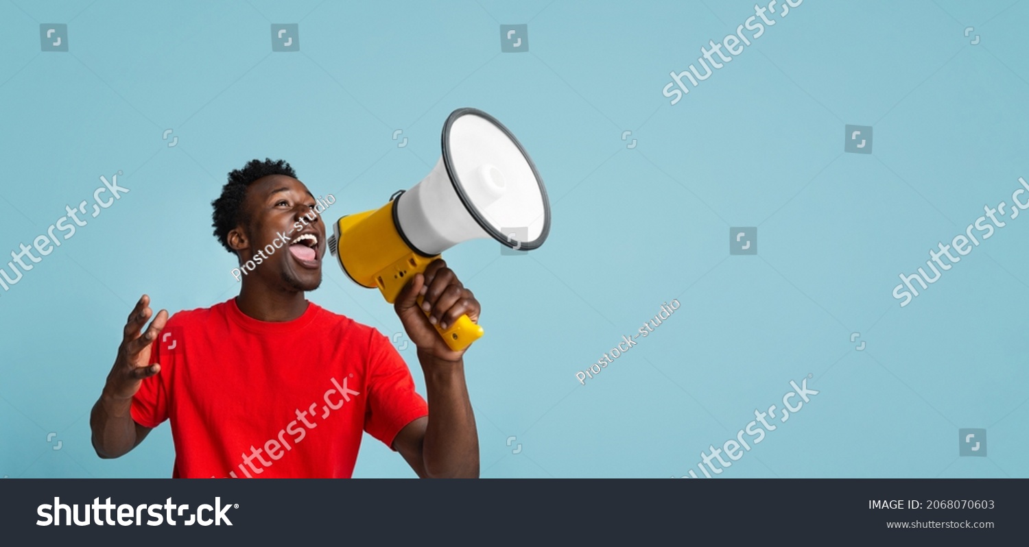 Emotional Young Black Guy Making Announcement With Megaphone In Hands, Cheerful African American Man Using Loudspeaker For Sharing News While Standing Over Blue Background, Panorama With Copy Space #2068070603