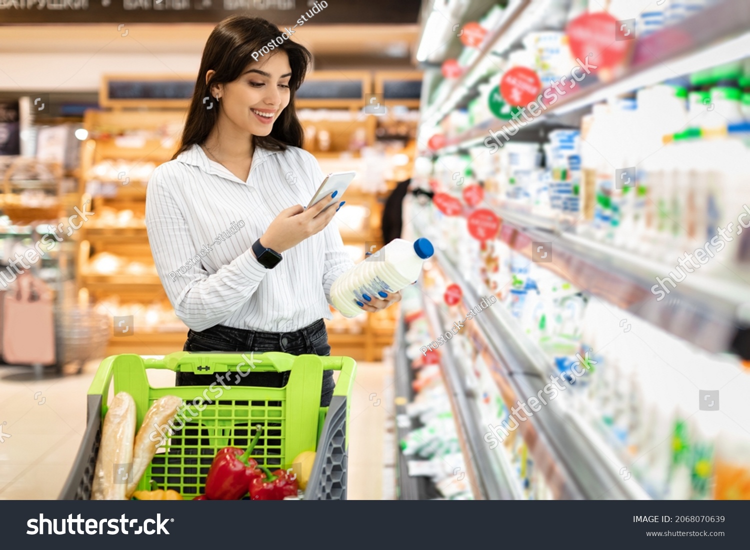 Middle Eastern Woman Using Phone Scanning Milk Bottle Doing Grocery Shopping In Supermarket, Standing With Shop Cart Full Of Products. Consumerism And Technologies, Customer's Application #2068070639