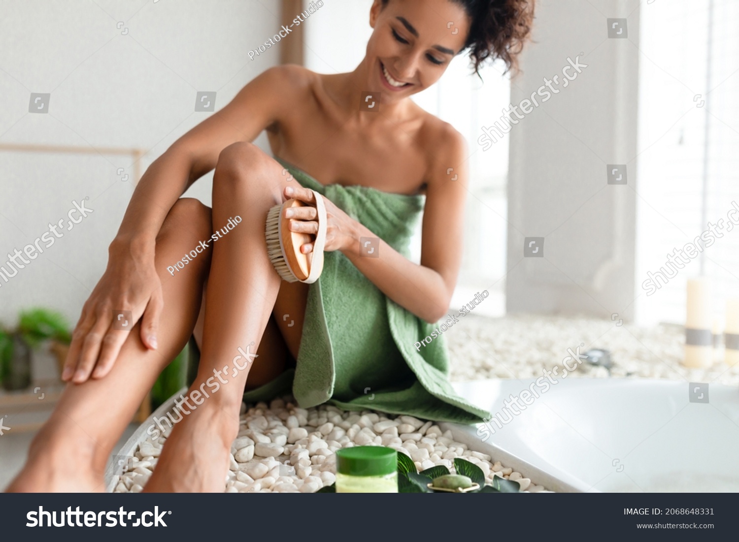 Young woman in towel making anti cellulite or lymphatic drainage massage near foamy bath at home, selective focus. Millennial lady dry brushing her leg, exfoliating skin, pampering herself in bathroom #2068648331