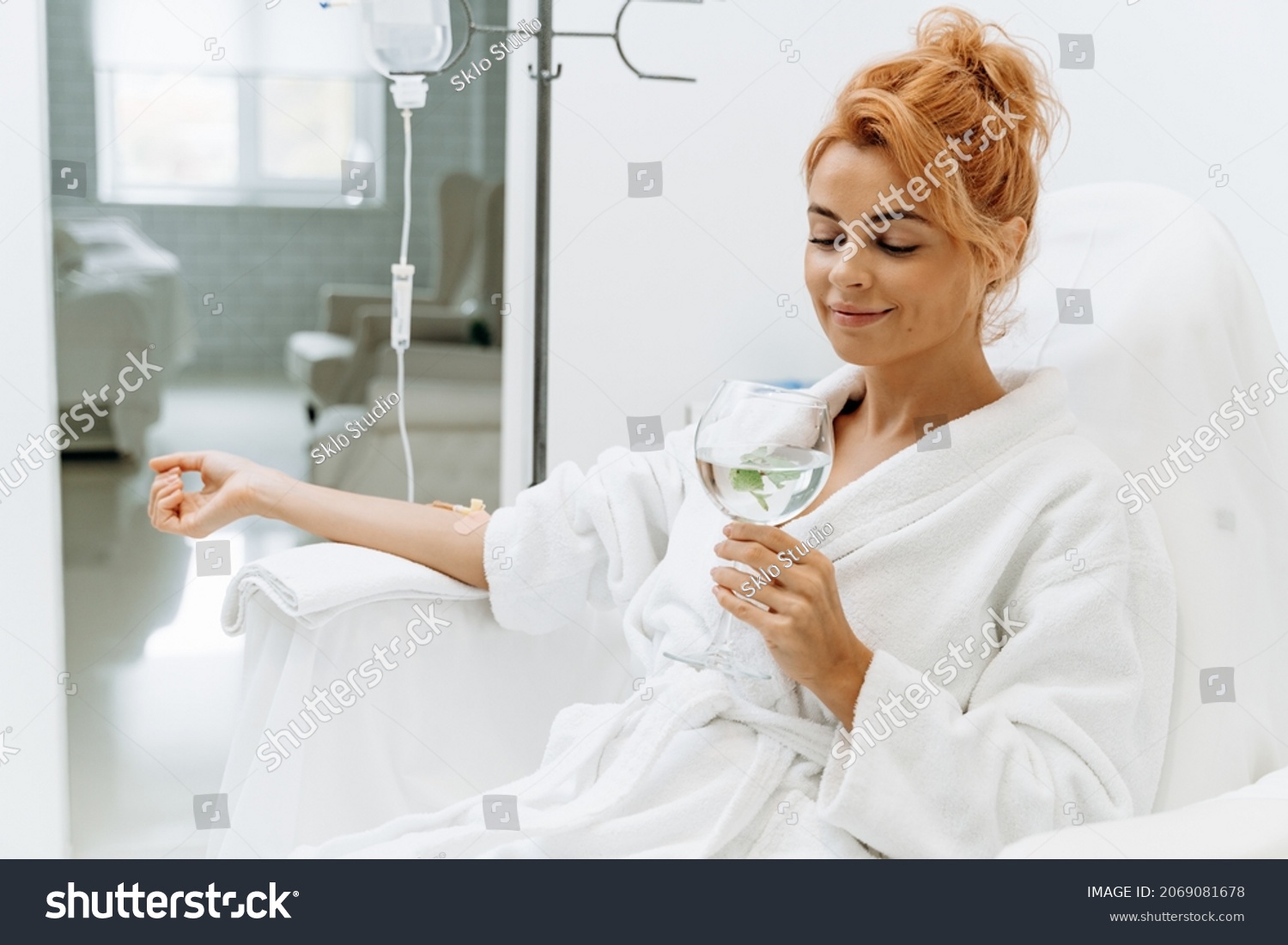 Waist up portrait view of the charming woman in white bathrobe sitting in armchair and receiving IV infusion. She is holding glass of lemon beverage and smiling  #2069081678