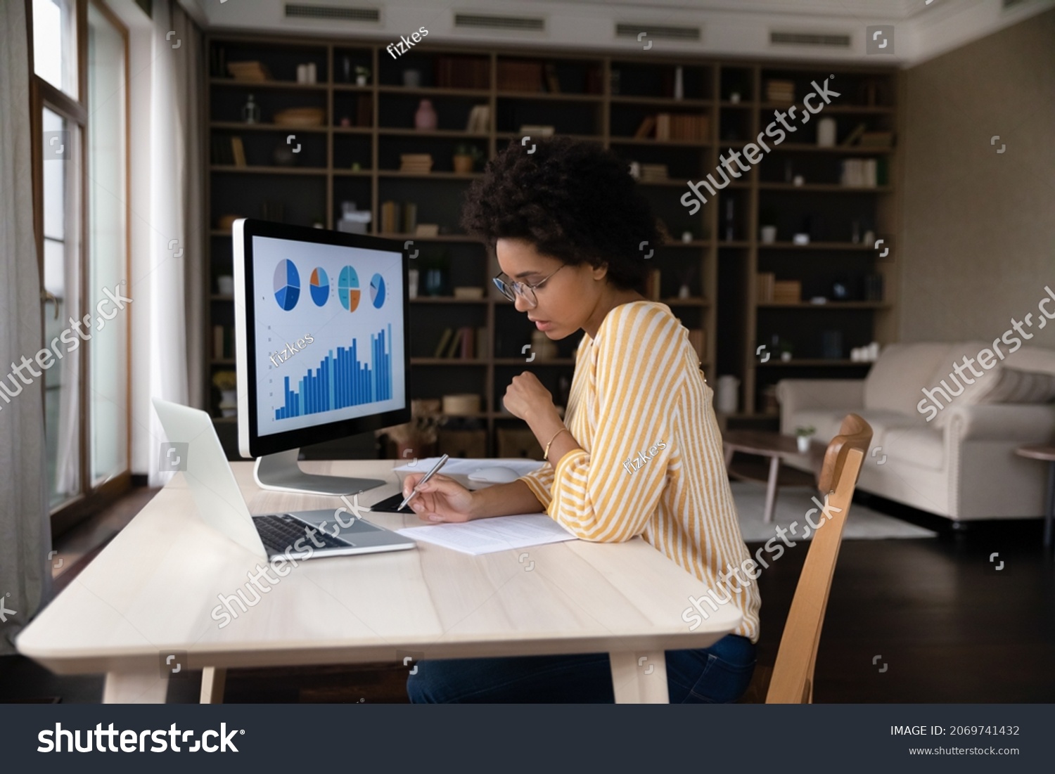 Concentrated young African American businesswoman employee worker in eyeglasses analyzing online sales statistics data on computer, reviewing marketing research, working with documents at home office. #2069741432