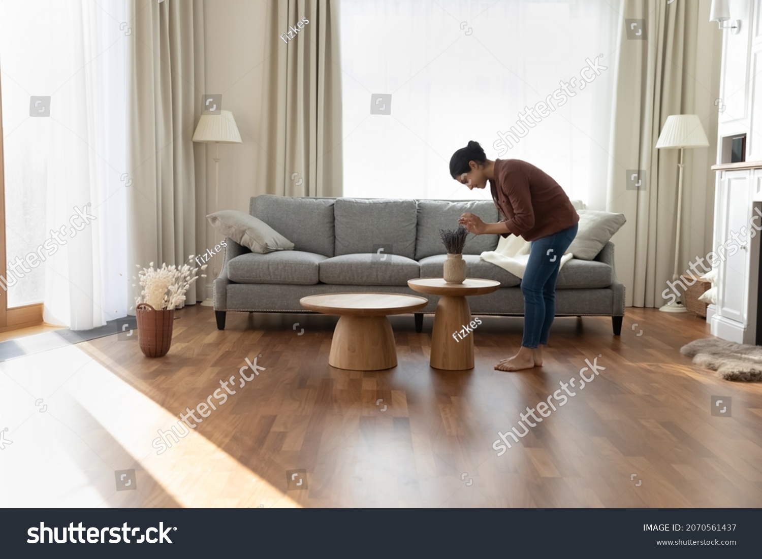 Full length of young Indian ethnicity housewife standing in modern spacious living room create cosiness at home, arranging flowers bunch in vase. Housework routine, interior designer workflow concept #2070561437