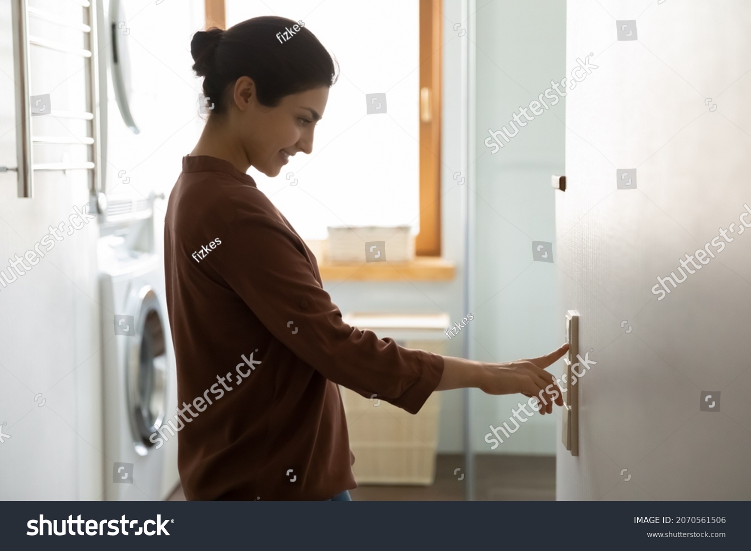 Young Indian ethnicity female push button turns off on light inside modern domestic laundry room, adjusting thermostat for controlling air conditioner and heater inside house. Climate control concept #2070561506