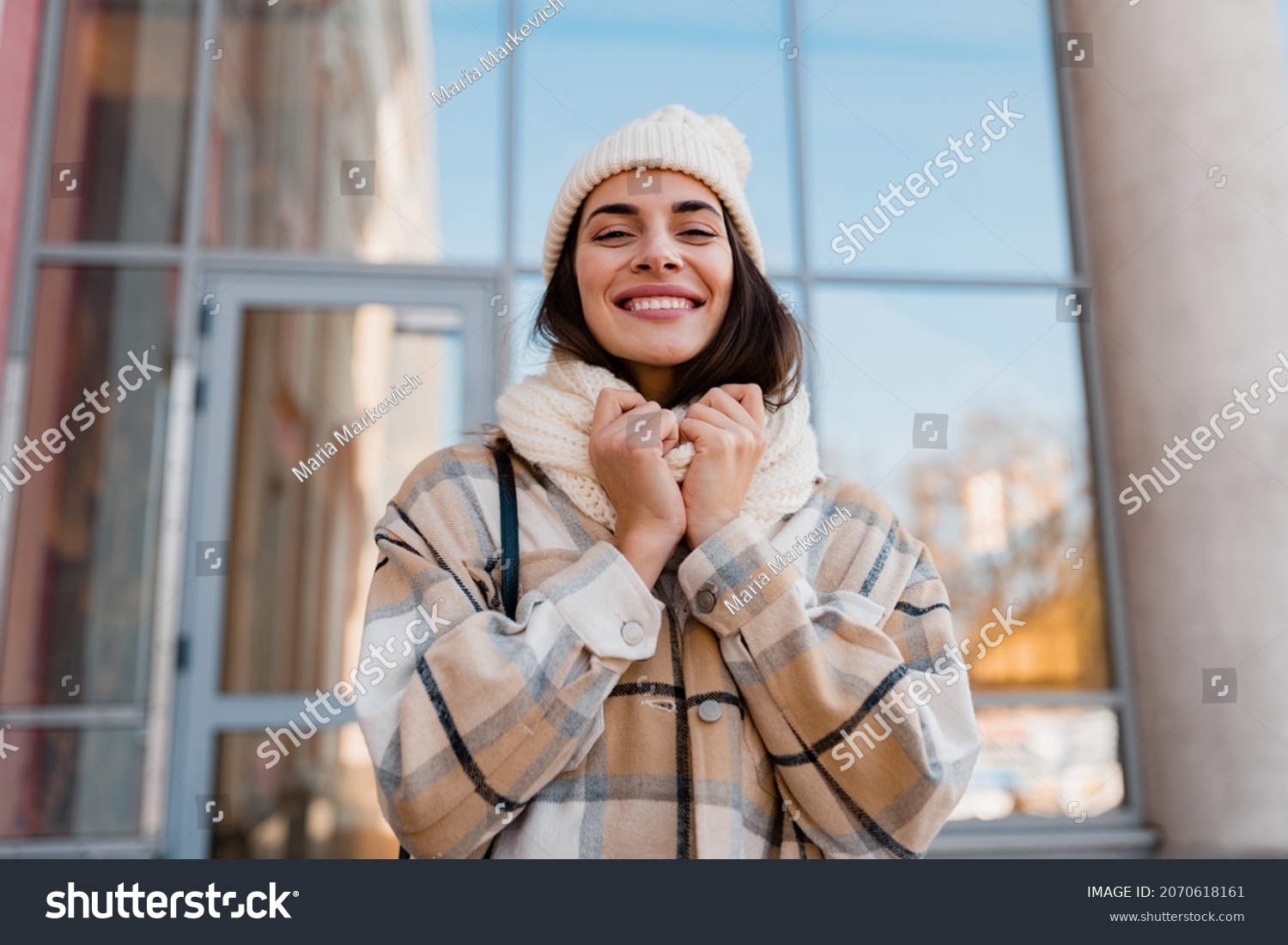 stylish attractive young smiling woman walking in street in winter outfit with coffee wearing checkered coat, white knitted hat and scarf, happy mood, fashion style trend #2070618161