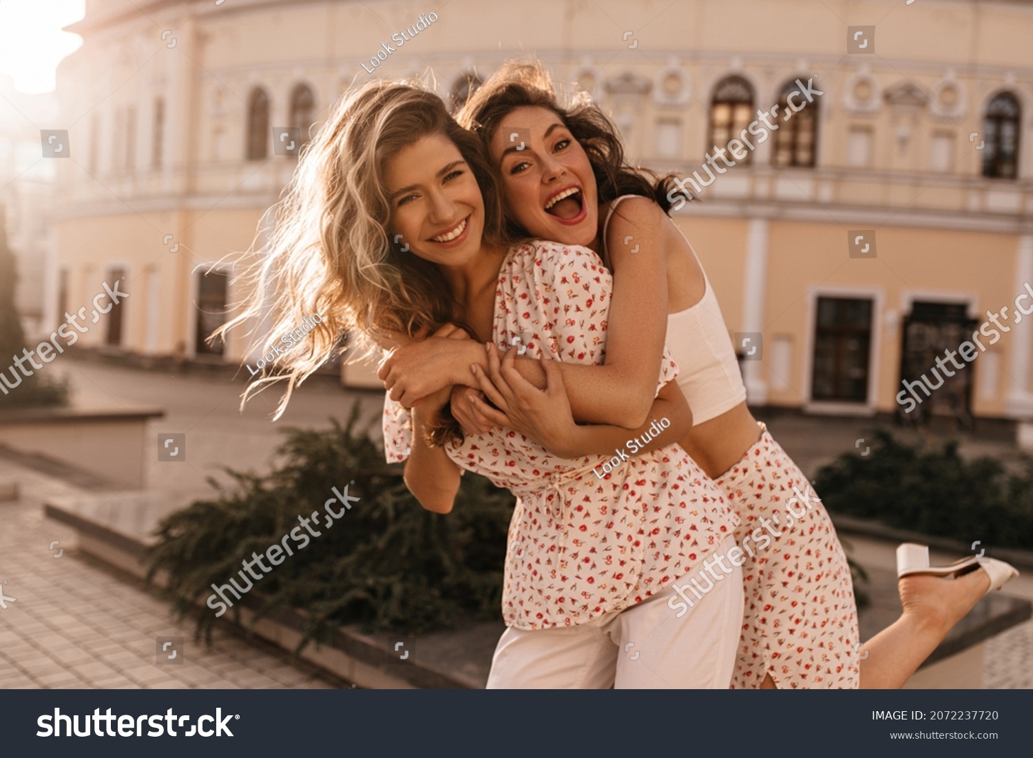 Caucasian young woman brunette hung on back of her friend against the open background of city. Girls are smiling broadly with their teeth at camera, dressed in white outfits. #2072237720