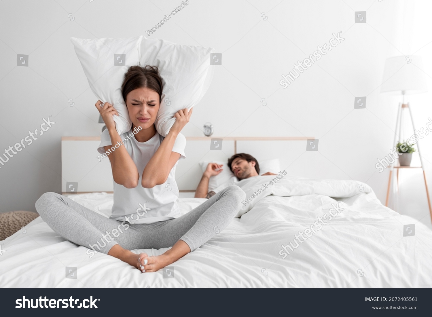 Stressed sad millennial caucasian lady sits on bed covers ears with pillow and suffers from snoring of sleeping man at night, empty space. Insomnia, sleep problems, apnea and relationship difficulties #2072405561