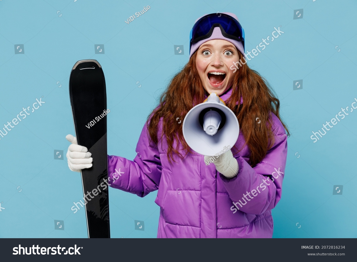 Skier woman in warm purple windbreaker jacket ski goggles mask spend extreme weekend in mountains hold scream in megaphone announces discounts sale Hurry up isolated on plain blue background studio. #2072816234